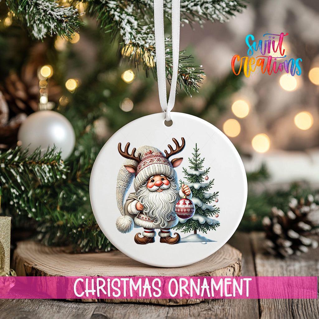 A white Christmas ornament featuring a Santa Claus figure holding a Christmas ornament and a pine tree, with a blurred background of a decorated Christmas tree and twinkling lights.