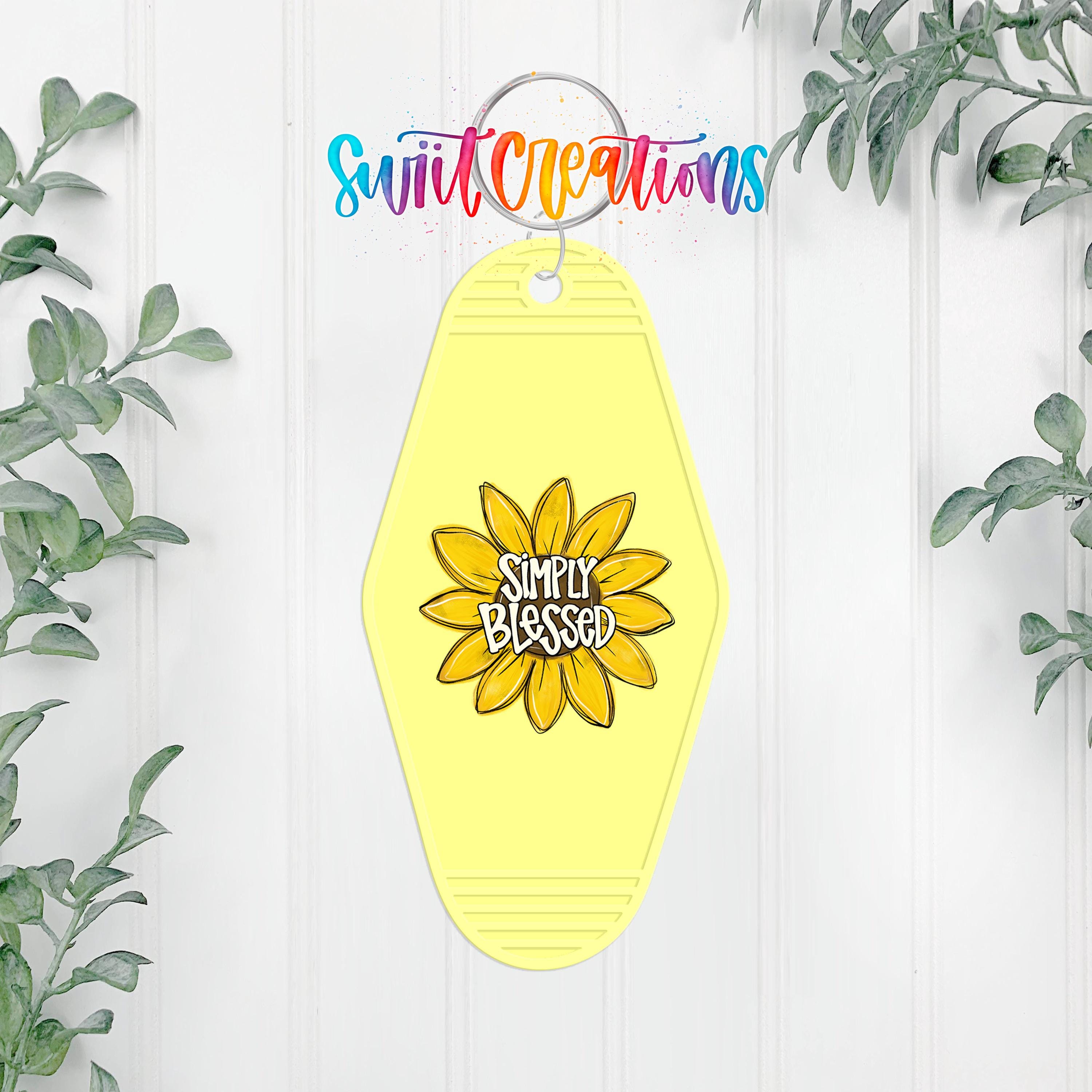 A yellow keychain with a flower design and the text "Simply Blessed" on it, hanging on a white wall with greenery in the background.