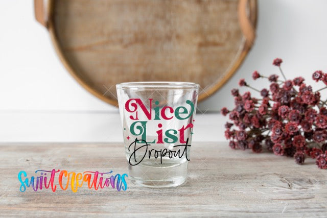 A shot glass with the text " Nice List Dropout" printed on it, placed on a wooden surface next to a bunch of red flowers.