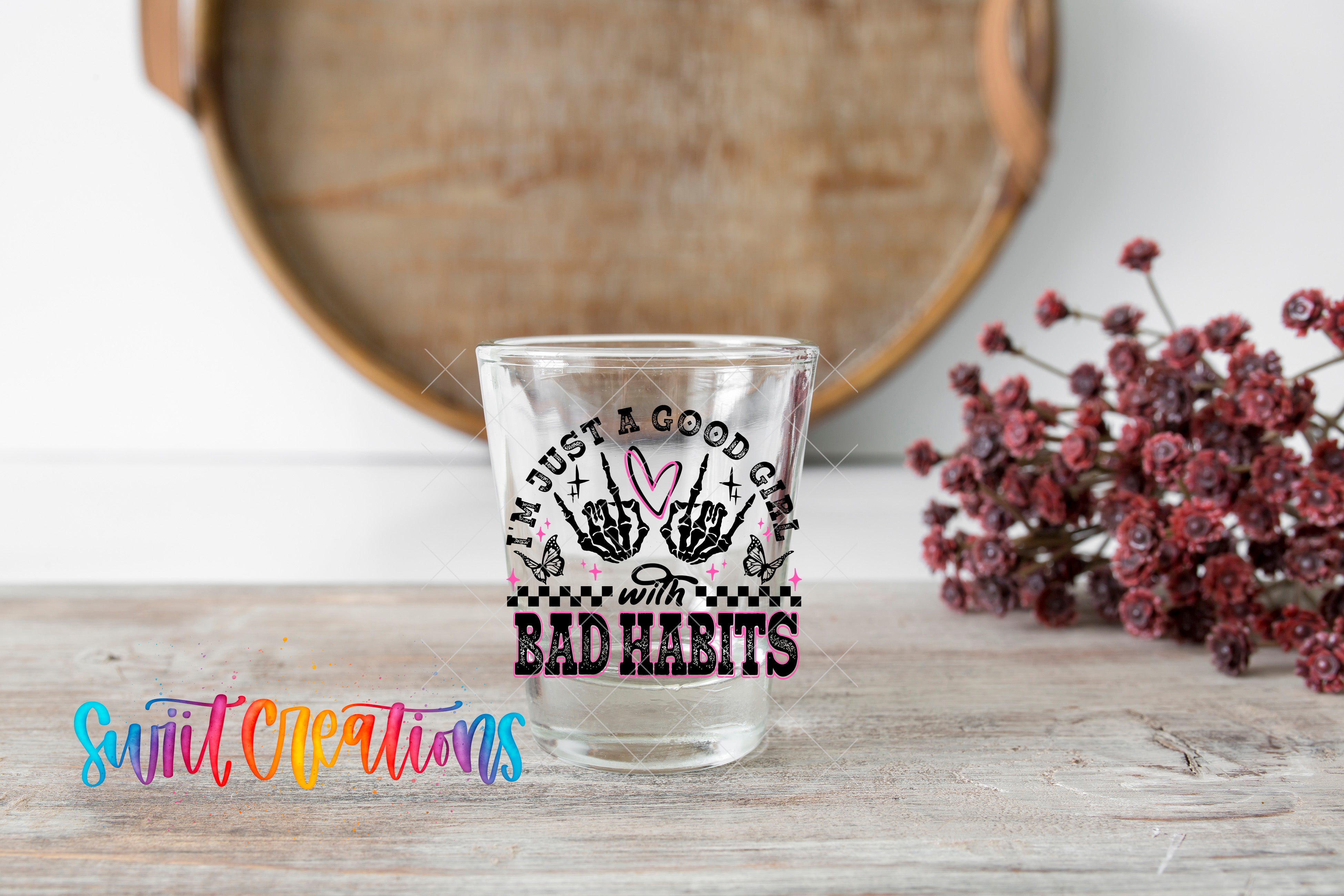 A clear glass with a hand-drawn design and text on it, placed on a wooden surface next to a bunch of red flowers.