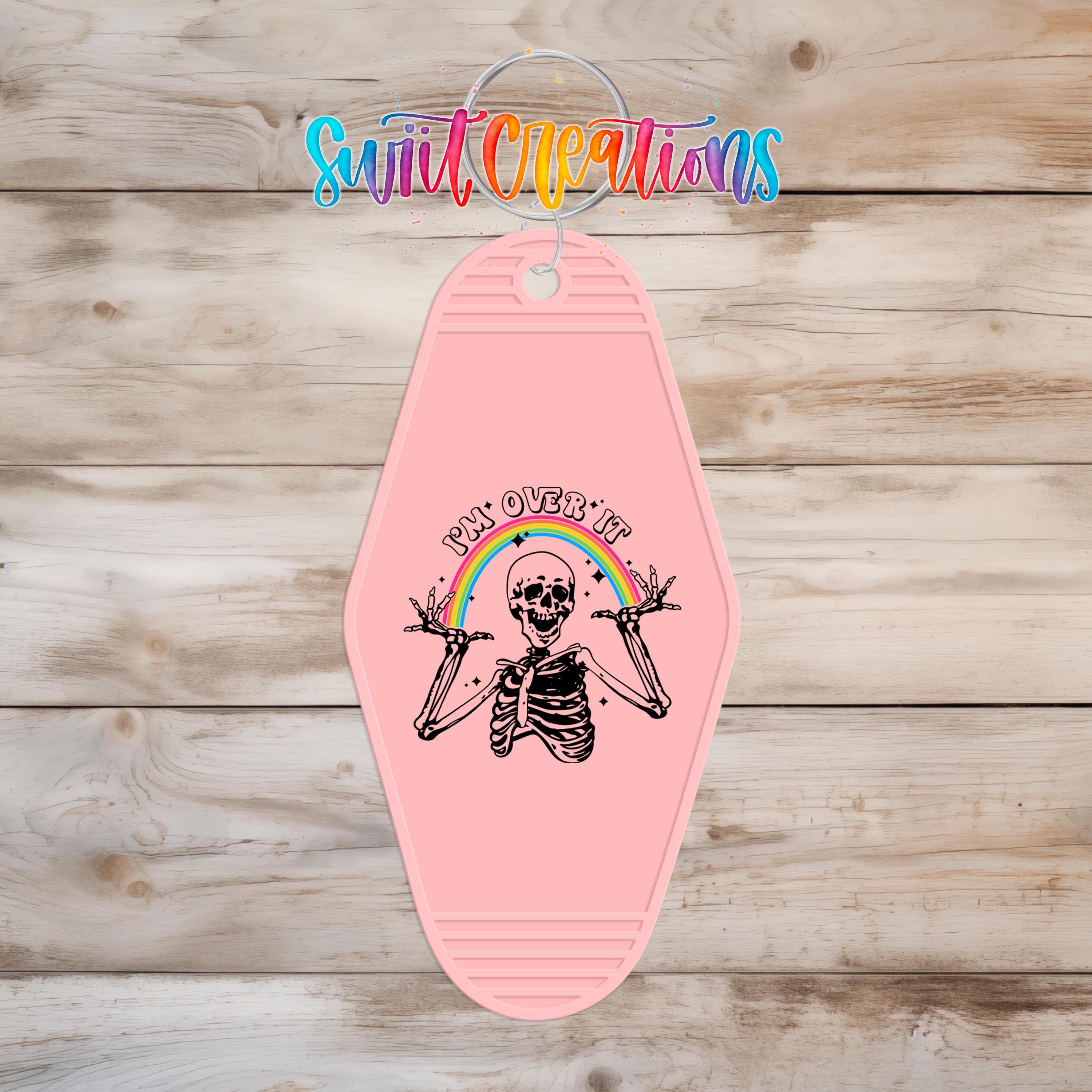 A pink keychain with a skeleton graphic and the text "My Over It" printed on it, hanging on a wooden surface.