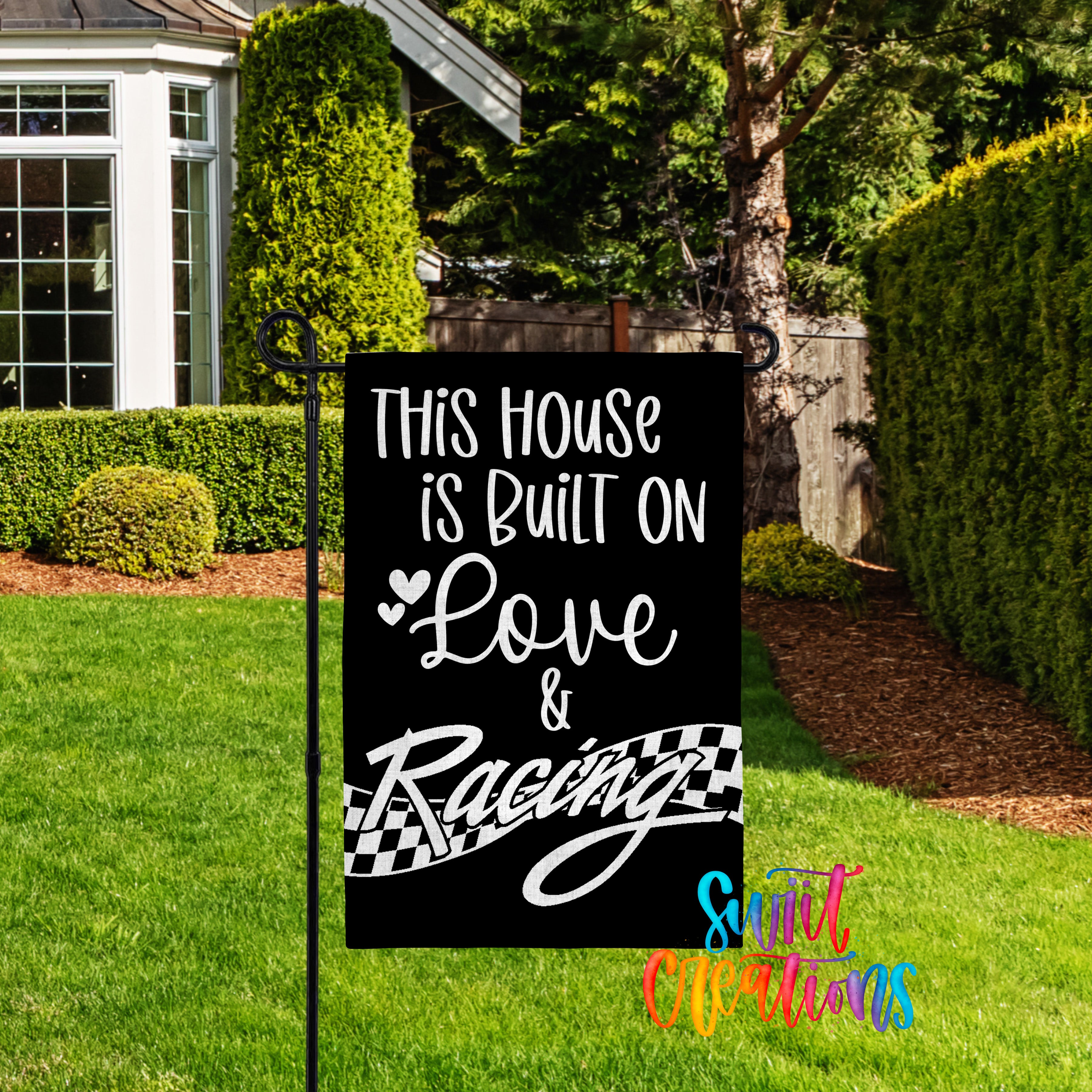 a black sign with white text that reads "This house is built on love & racing" in a grassy yard with a house and trees in the background.