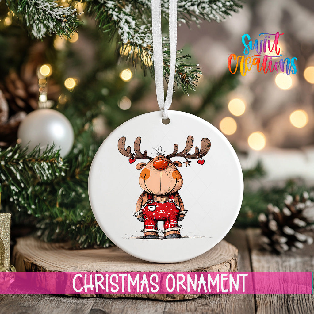 A white Christmas ornament featuring a cartoon reindeer is hanging from a ribbon, with a blurred background of a decorated Christmas tree and other ornaments.