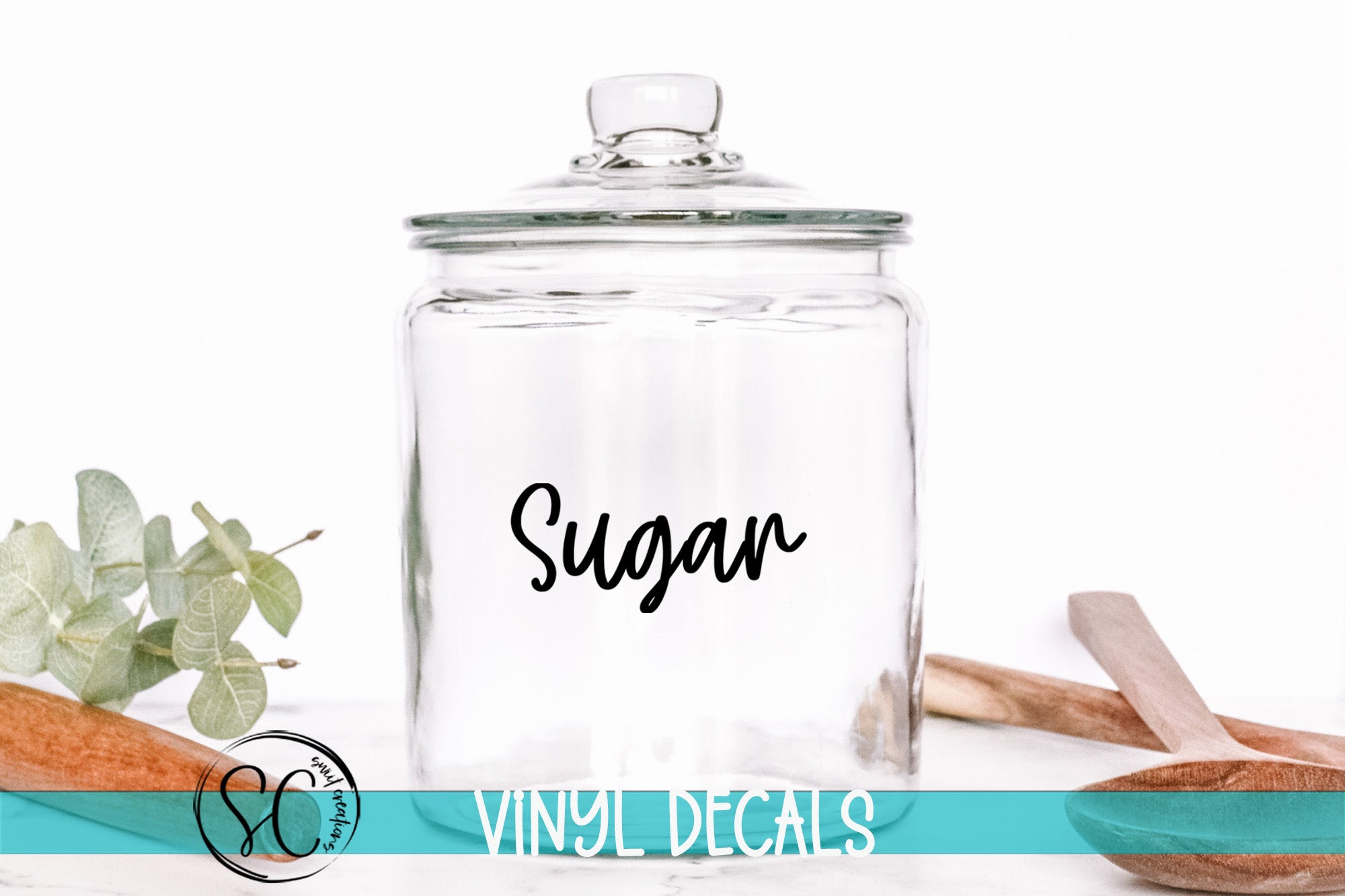 A clear glass jar with the word "Sugar" written on it, placed on a white surface next to some wooden utensils.
