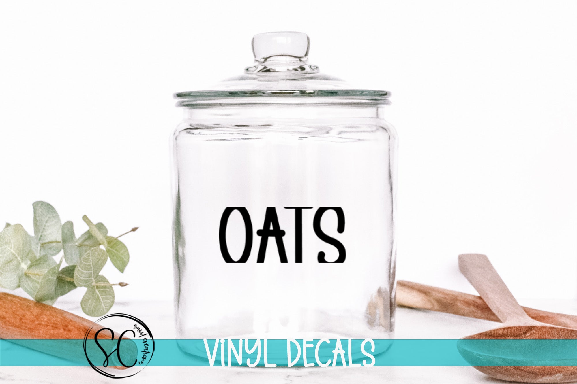 A clear glass jar with the word "OATS" printed on it, placed on a white surface next to some wooden utensils.