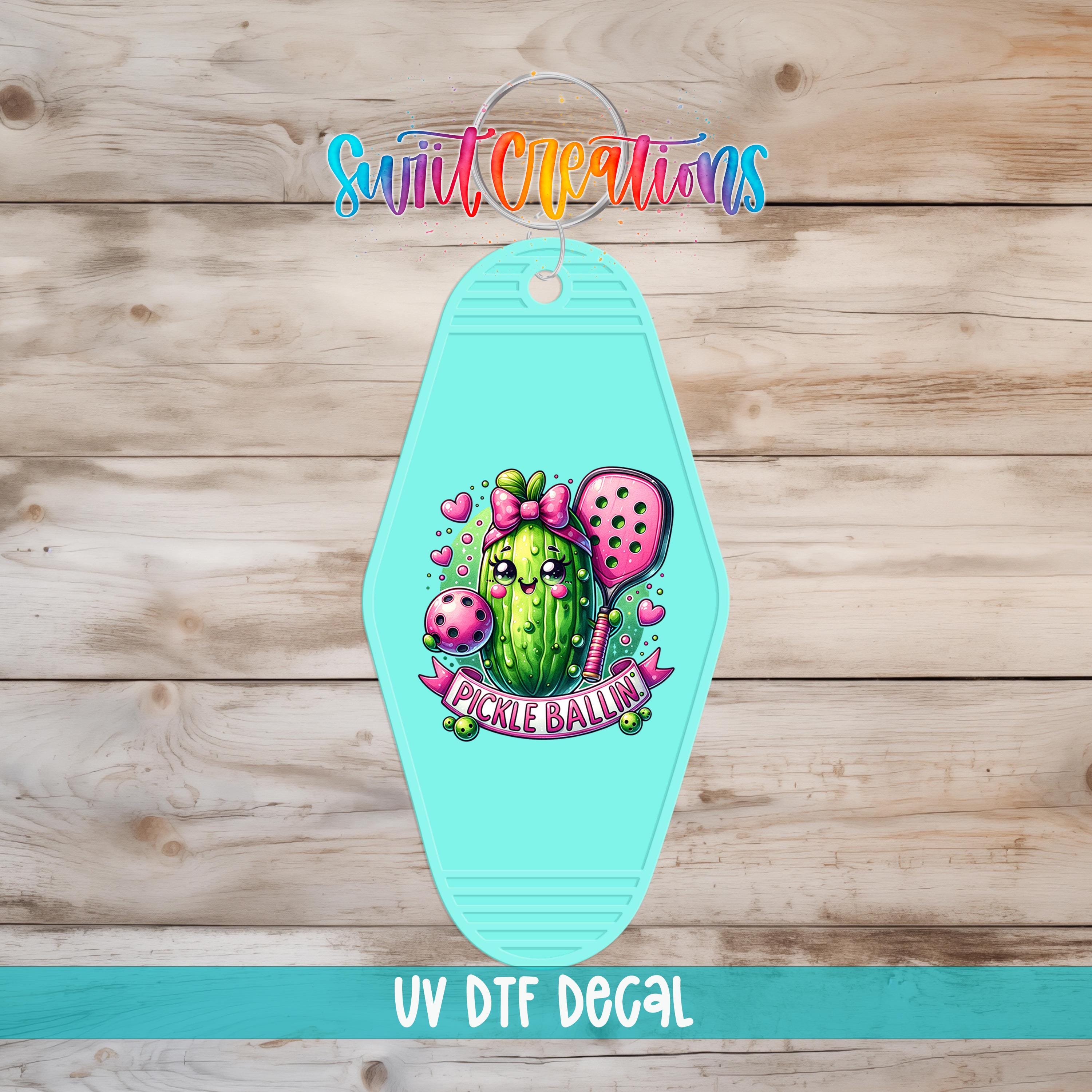 A turquoise keychain with a cartoon image of a pickle ball and a bow on it, along with the text "Pickle Ballin'" and "Swift Creations".
