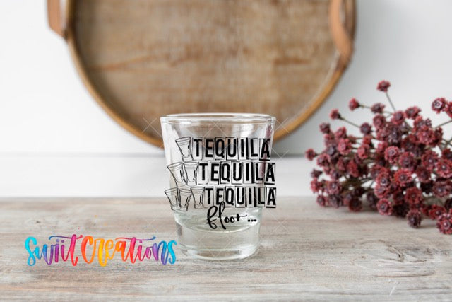 A shot glass with the text "TEQUILA TEQUILA TEQUILA fleet..." printed on it, placed on a wooden surface next to a bunch of red flowers.