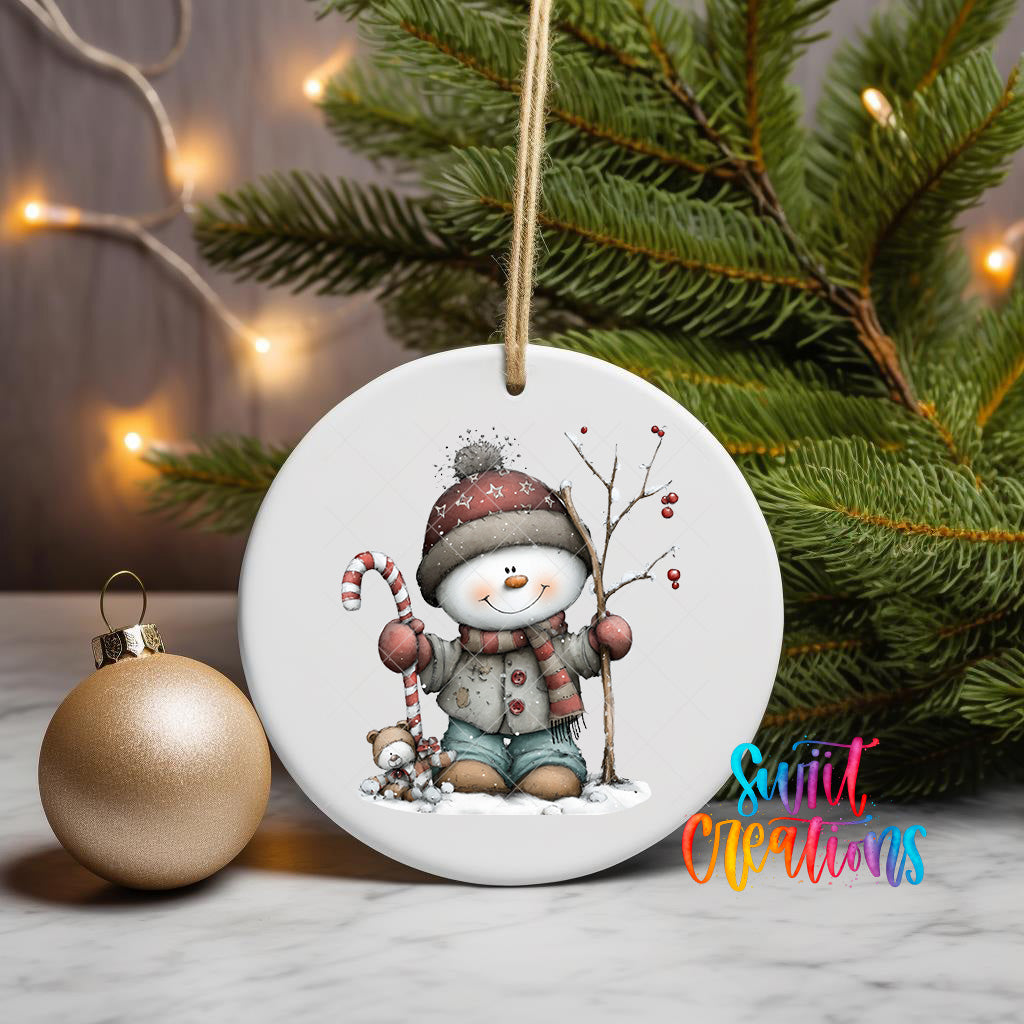 A white ceramic ornament featuring a snowman and a candy cane, placed on a marble surface next to a gold ornament and a Christmas tree.