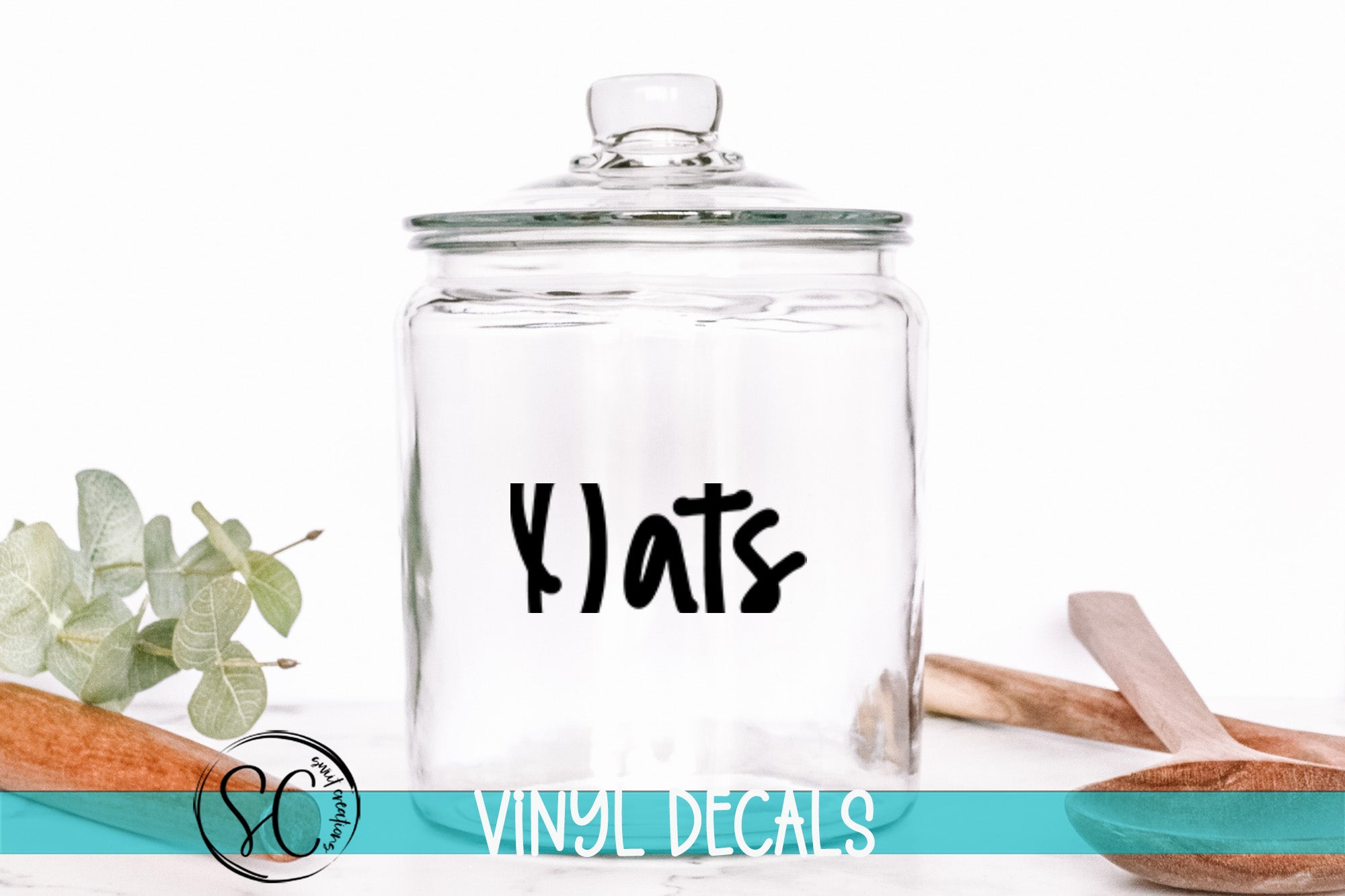A clear glass jar with a black label that says "Y'ats" is placed on a white surface, surrounded by various kitchen utensils and a plant.