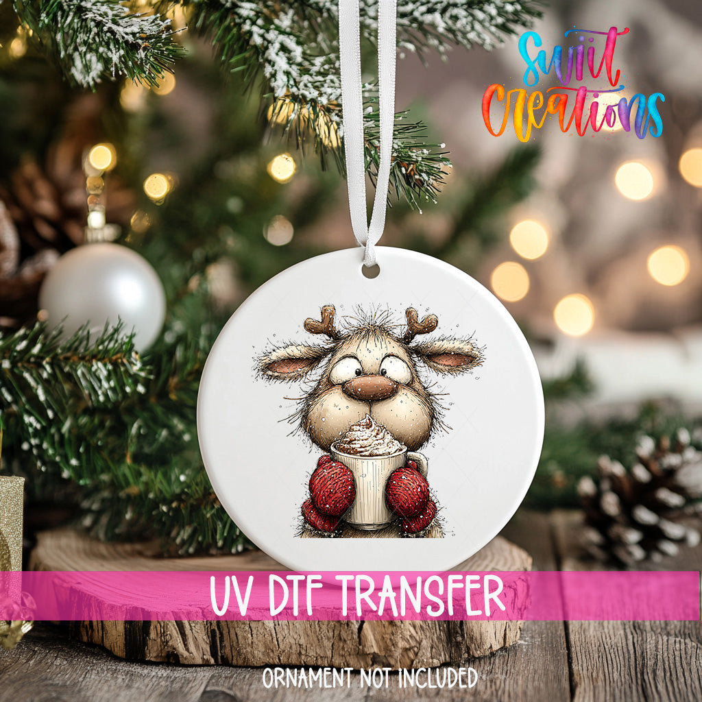 A white ornament with a cartoon reindeer holding a cup of hot chocolate is hanging on a Christmas tree.