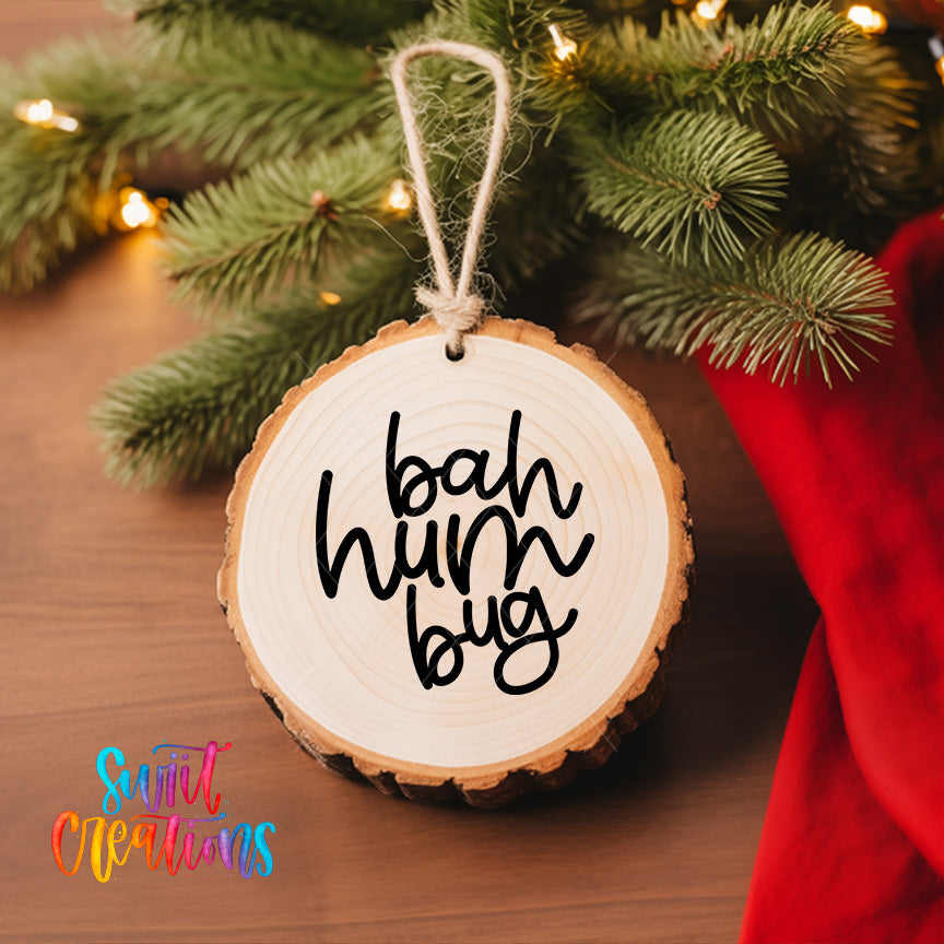 A wooden ornament with the text "bah hum bug" hanging from a tree branch, surrounded by pine branches and twinkling lights.