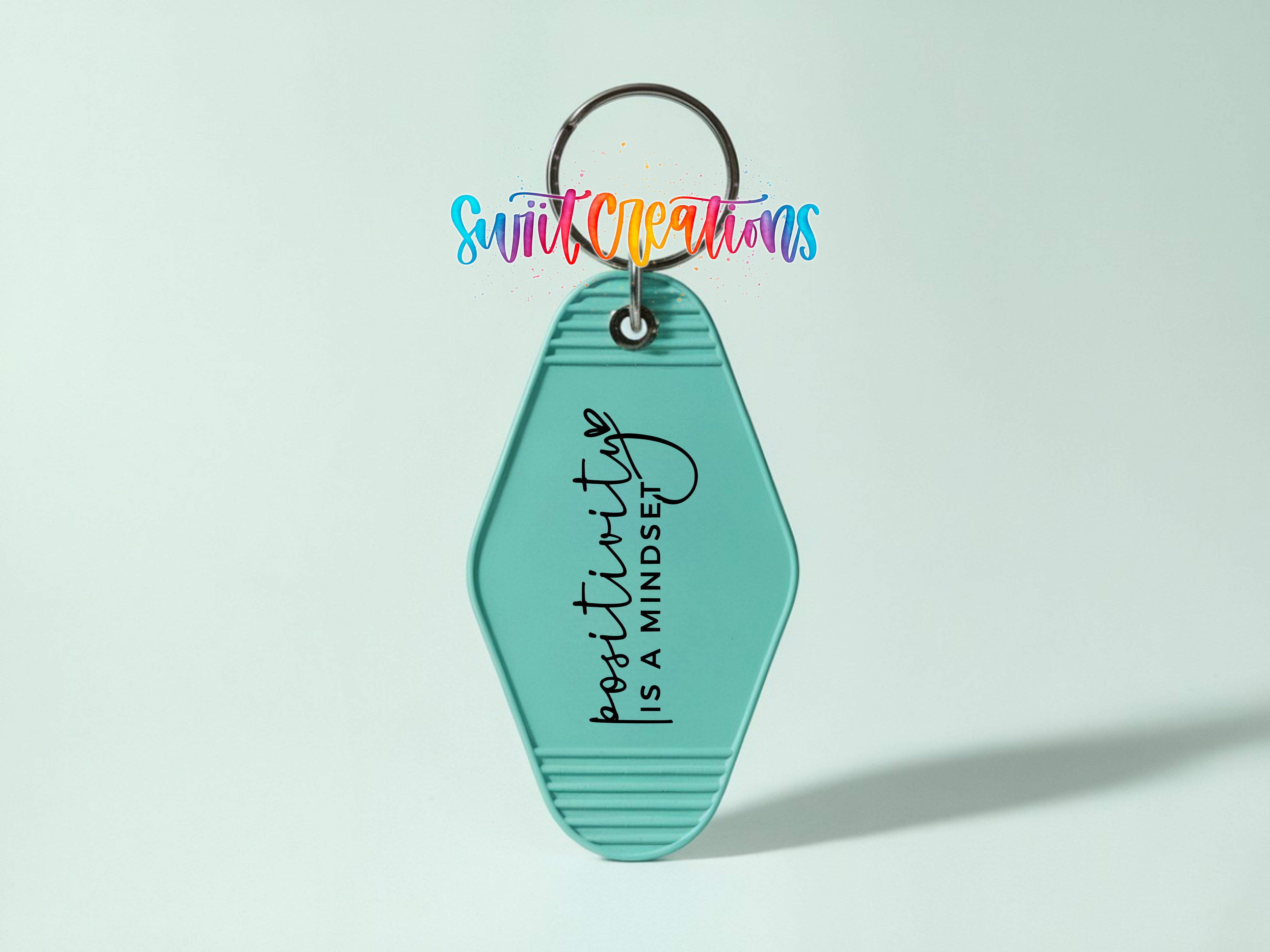 A turquoise keychain with the text "positivity is mindset" printed on it, along with a colorful, hand-lettered logo above it.