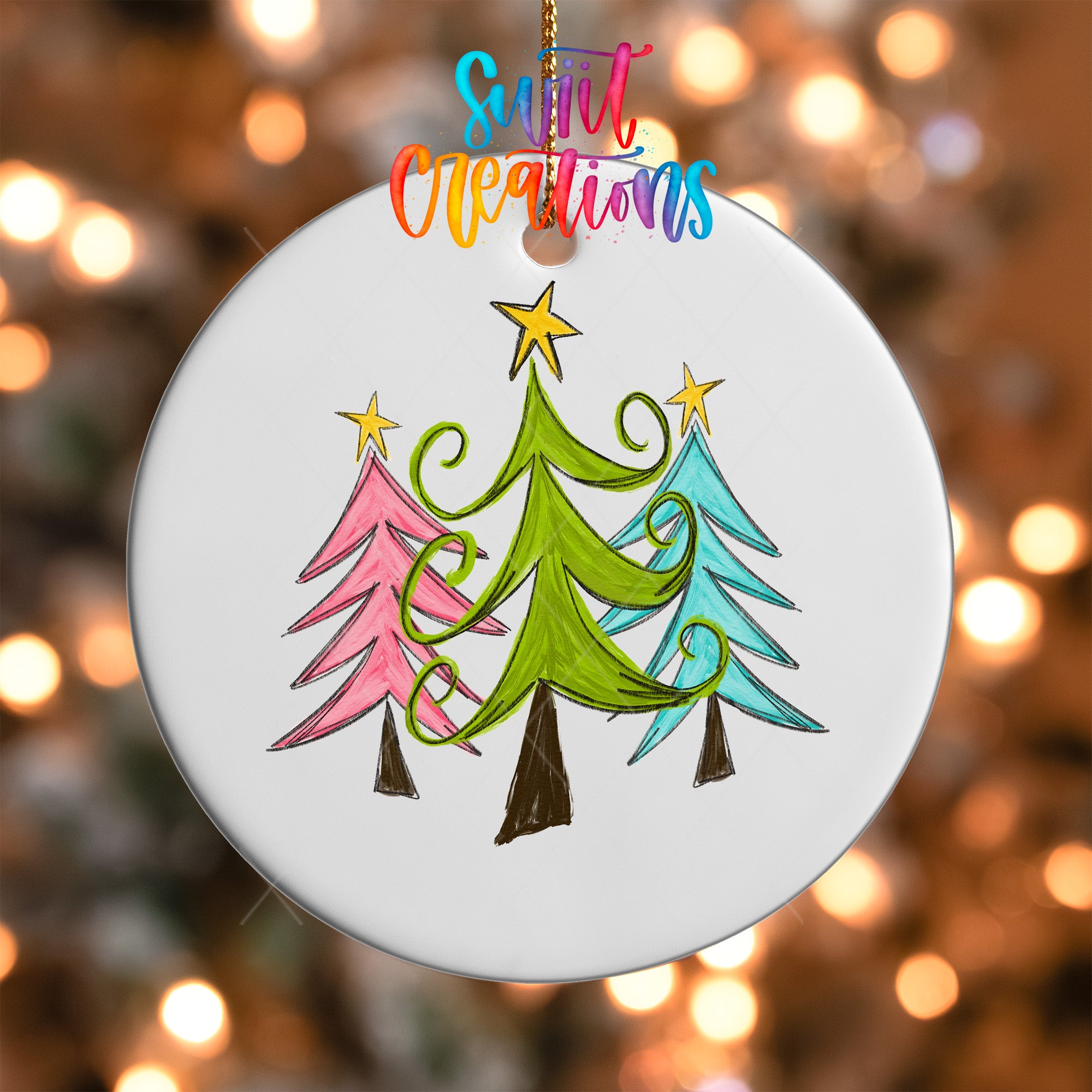 A white ornament with a drawing of three stylized Christmas trees in different colors, including green, pink, and blue, against a blurred background with colorful lights.