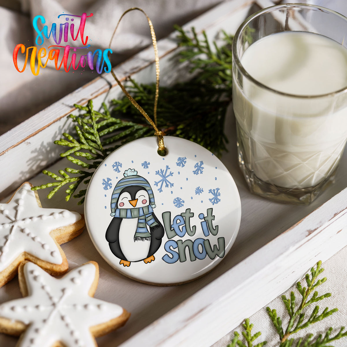 A white ornament with a penguin wearing a scarf and hat, holding a snowflake, and the text "Let it snow" is displayed on a white tray alongside a glass of milk and some cookies.