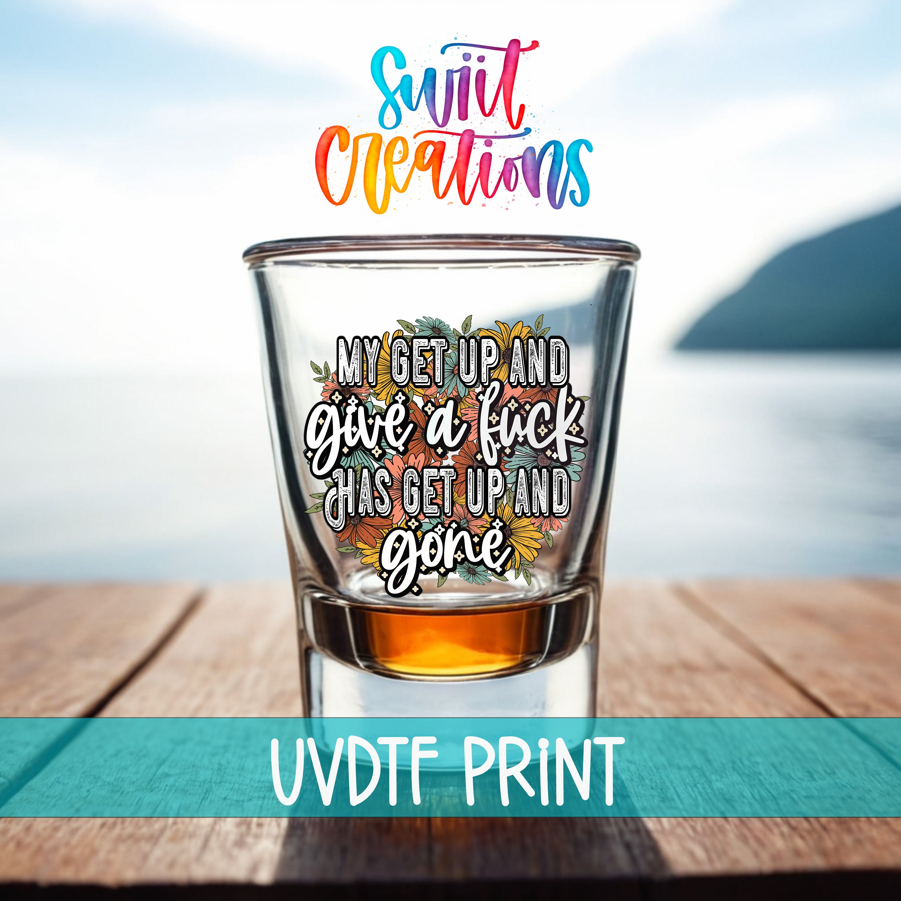 a shot glass with a colorful, artistic design and text on it, placed on a wooden surface with a body of water in the background.