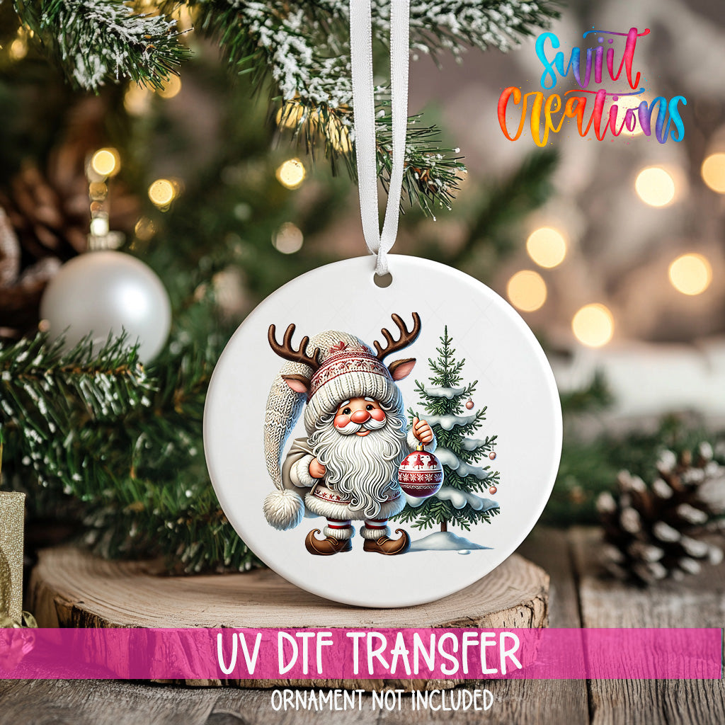 A white ornament featuring a Santa Claus figure holding a Christmas ornament and a pine tree, hanging on a Christmas tree.