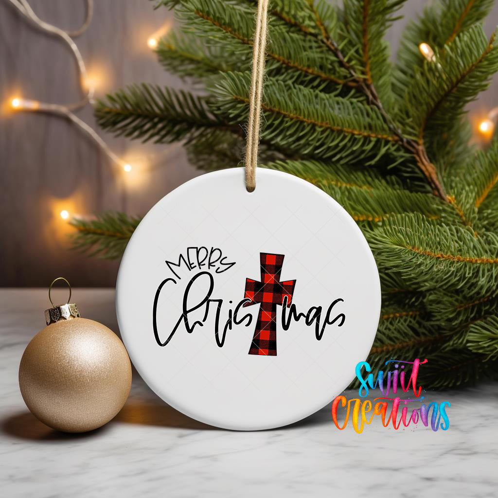 A white Christmas ornament with the text "Merry Christmas" and a red and black plaid cross is displayed on a table, with a gold ornament and a pine tree in the background.