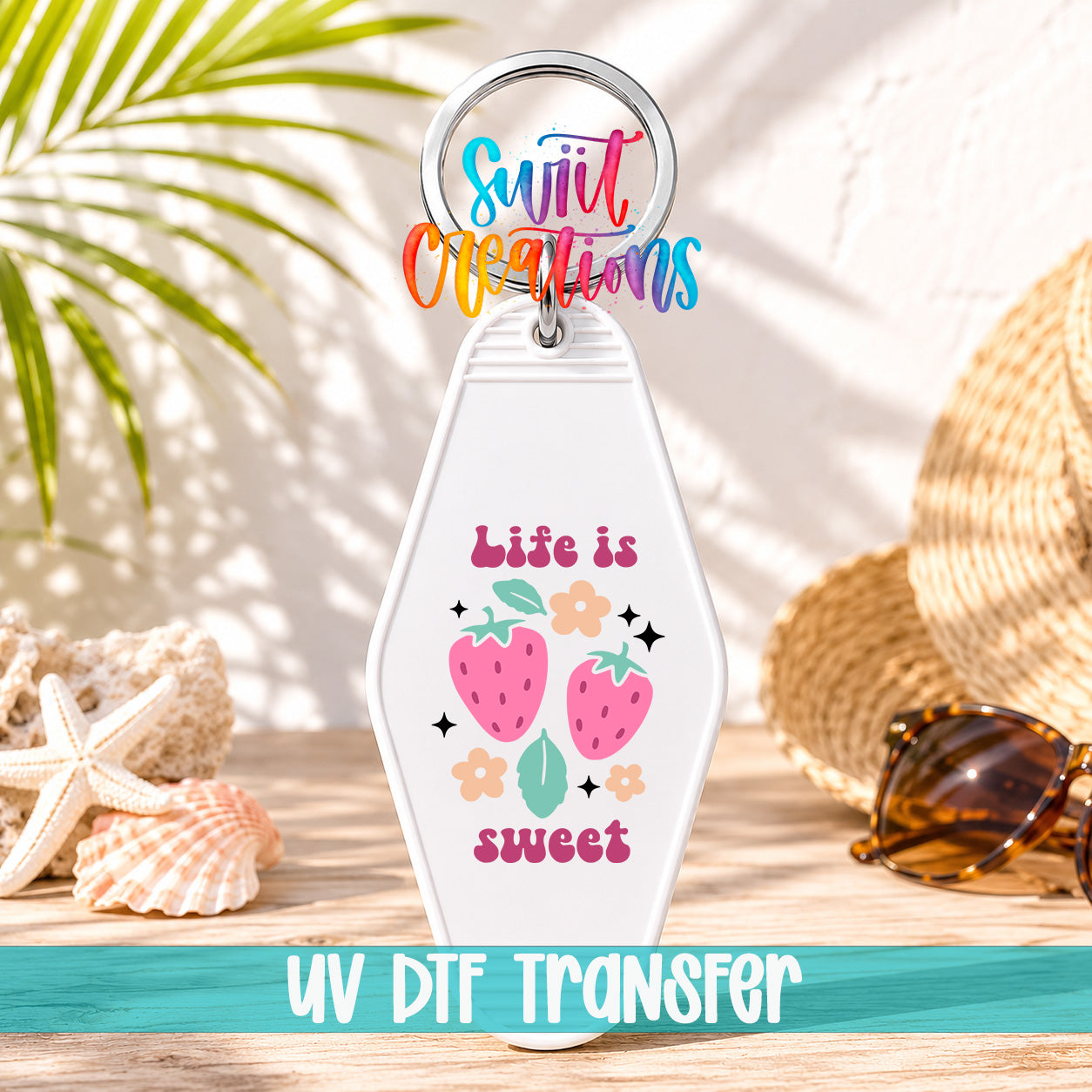 A keychain with a colorful design featuring strawberries and the text "Life is sweet" is displayed on a wooden surface, with sunglasses and a straw hat in the background.