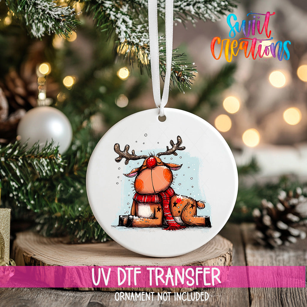 A white ceramic ornament with a cartoon reindeer design hangs on a Christmas tree, with a blurred background of pine branches and twinkling lights.
