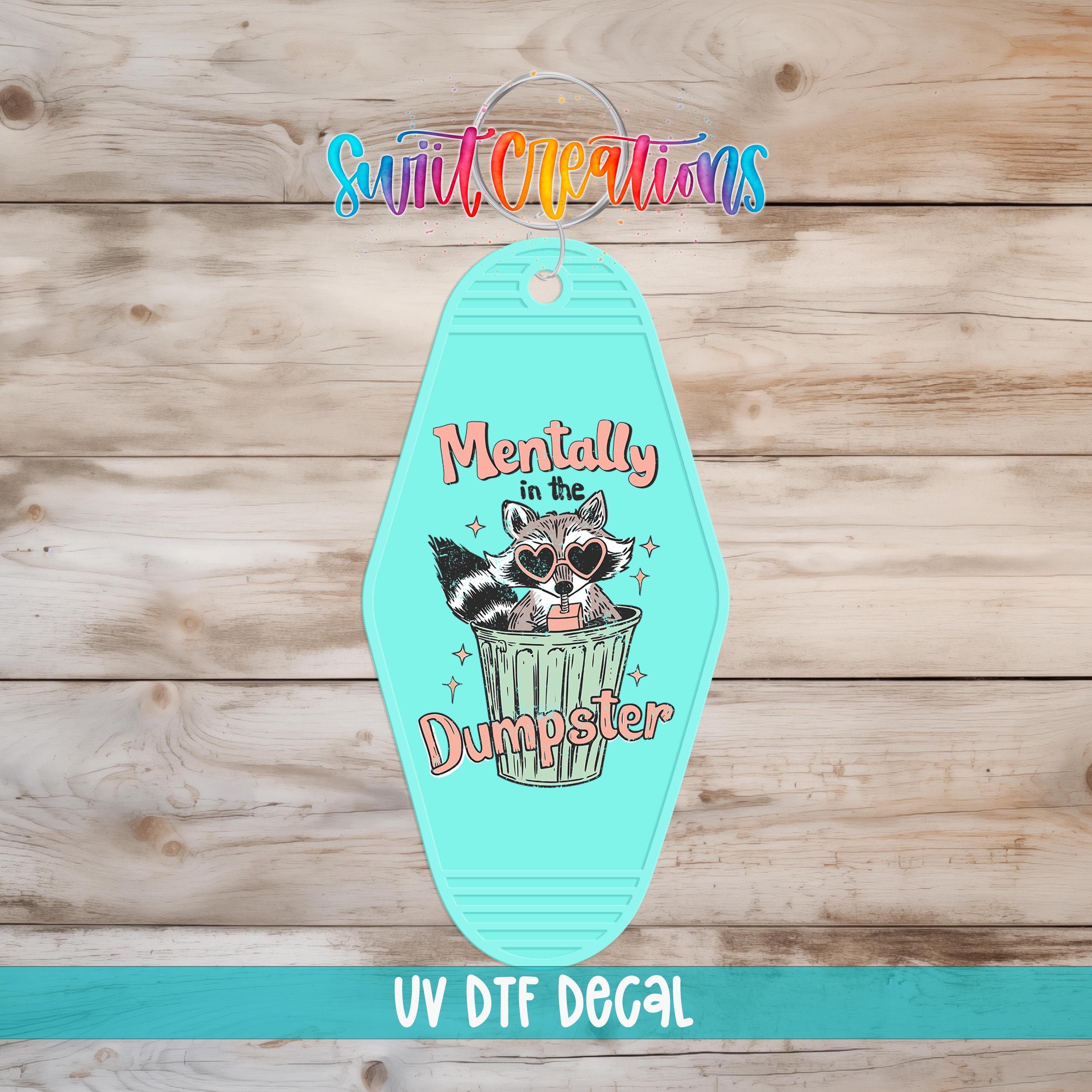 A turquoise keychain with a cartoon raccoon and the text "Mentally Dumpster" printed on it, hanging on a wooden surface.
