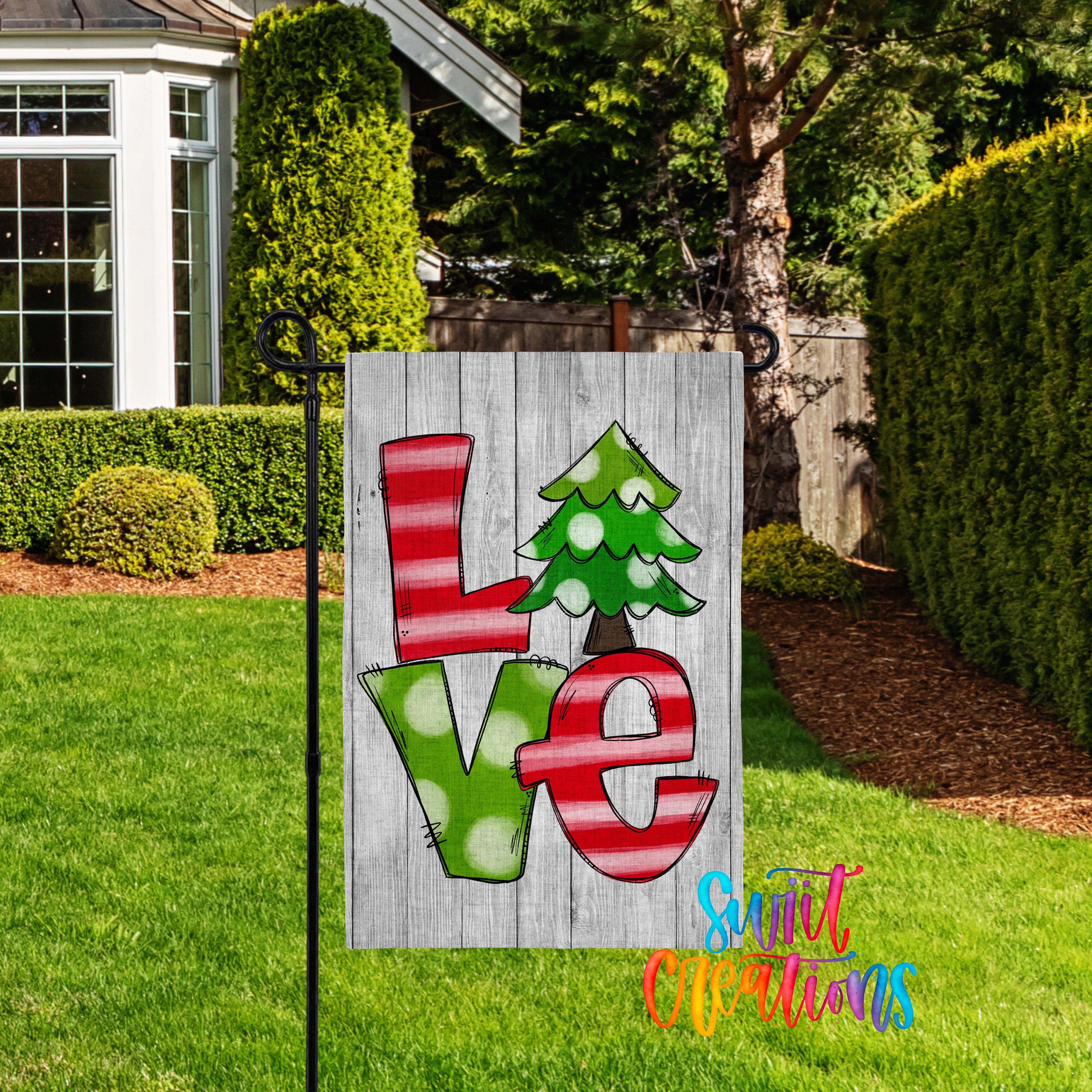 A wooden sign with the word "LOVE" painted on it, featuring a Christmas tree design, is displayed in a yard with a house and trees in the background.