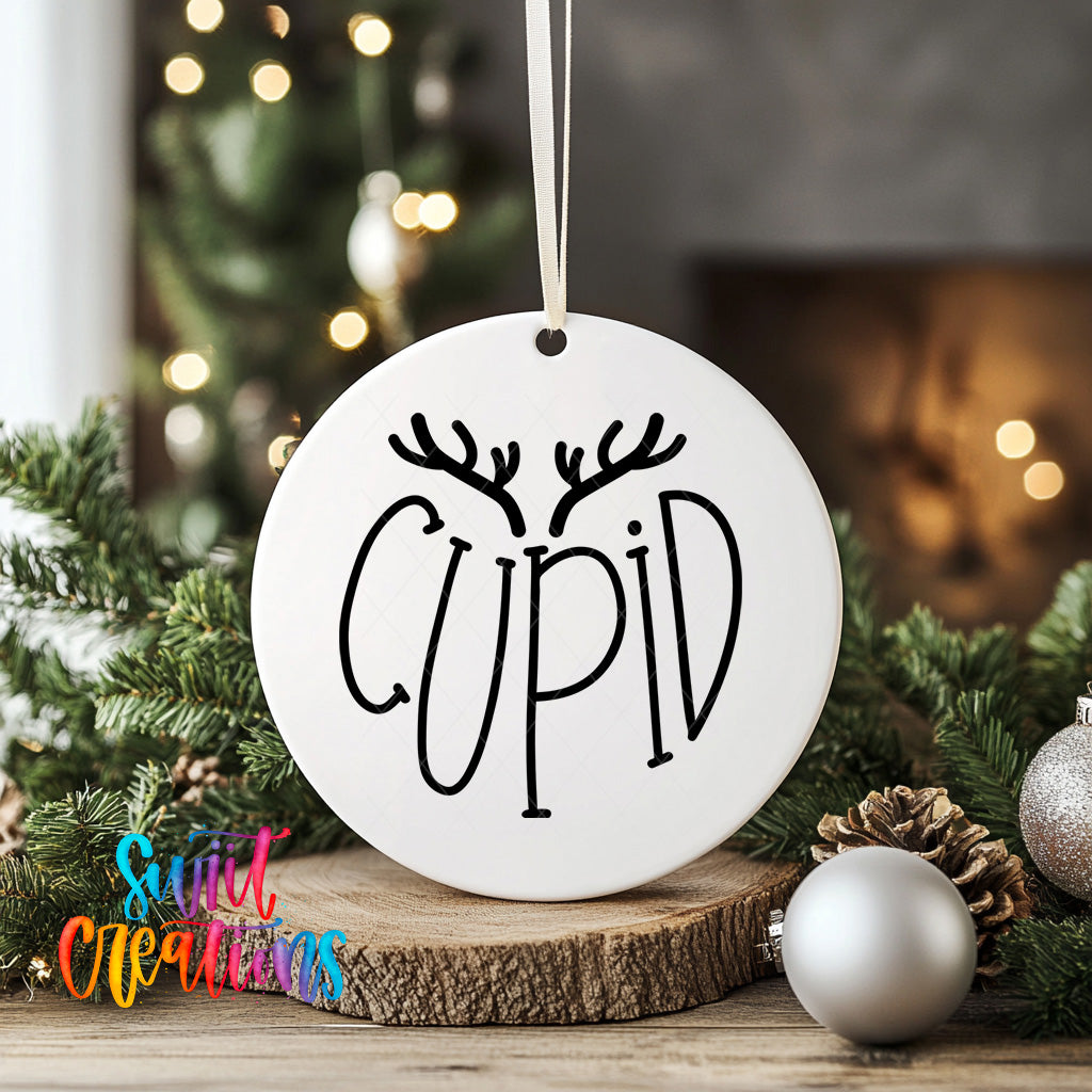 A white ornament with the word "CUPID" written on it, hanging from a string, surrounded by Christmas decorations such as pine branches and ornaments.