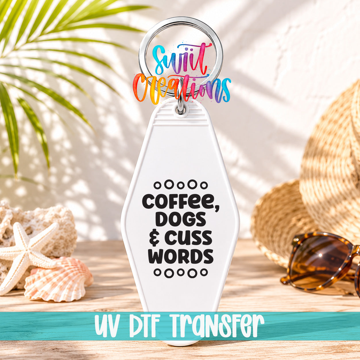 A keychain with the text "COFFEE, DOGS & CUSS WORDS" printed on it, along with a colorful ring and a straw hat. The keychain is placed on a wooden surface, with sunglasses and a seashell nearby.
