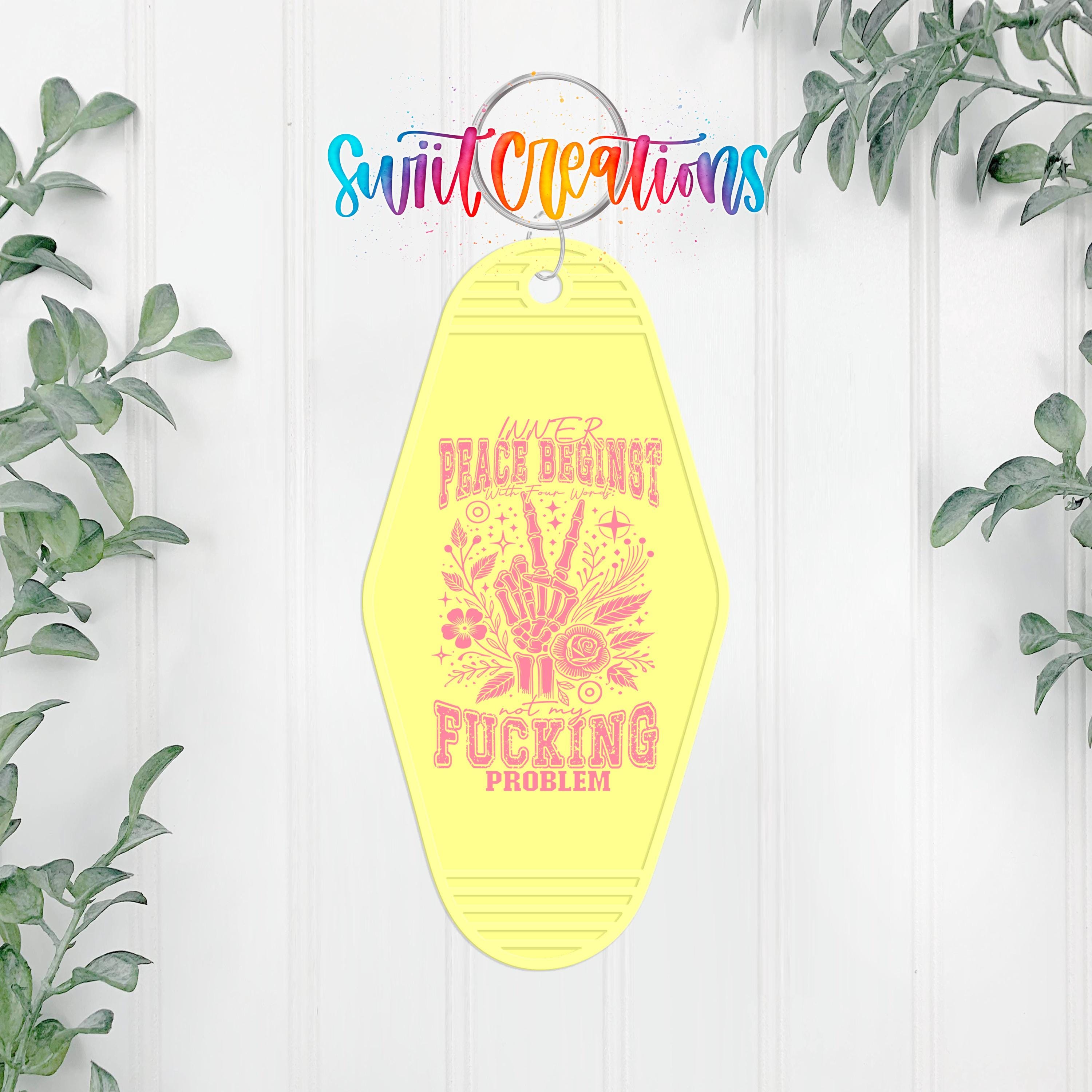 A yellow keychain with a pink and white design and text, hanging on a white wall with greenery in the background.