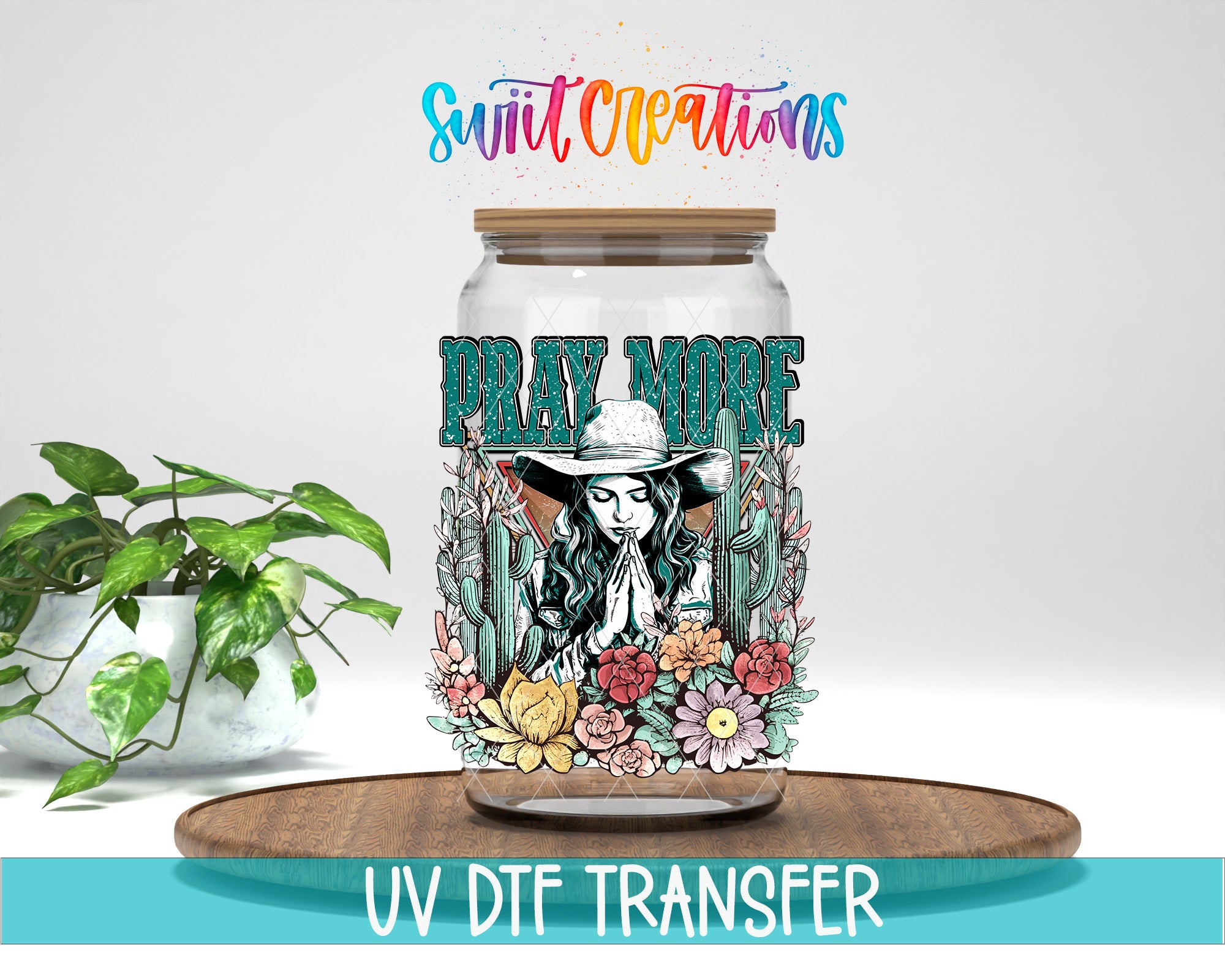 a glass jar with a colorful floral design and the text "PRAY MORE" printed on it. The jar is placed on a wooden surface, and there is a potted plant in the background.