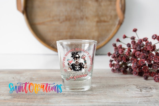 A shot glass with a graphic design featuring a woman and the word "Tequila" on it, placed on a wooden surface next to a bunch of red flowers.