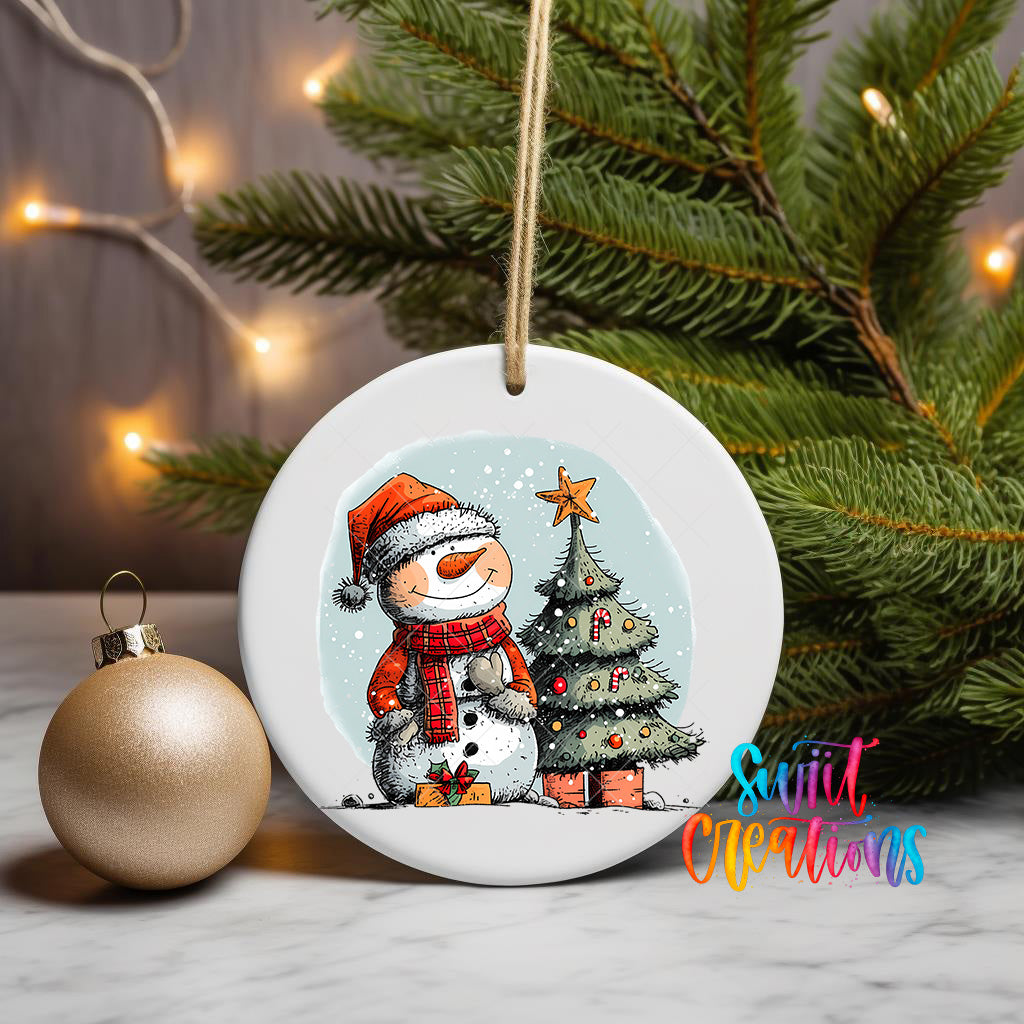 A white ceramic ornament featuring a snowman and a Christmas tree, with a gold ornament and a string of lights in the background.