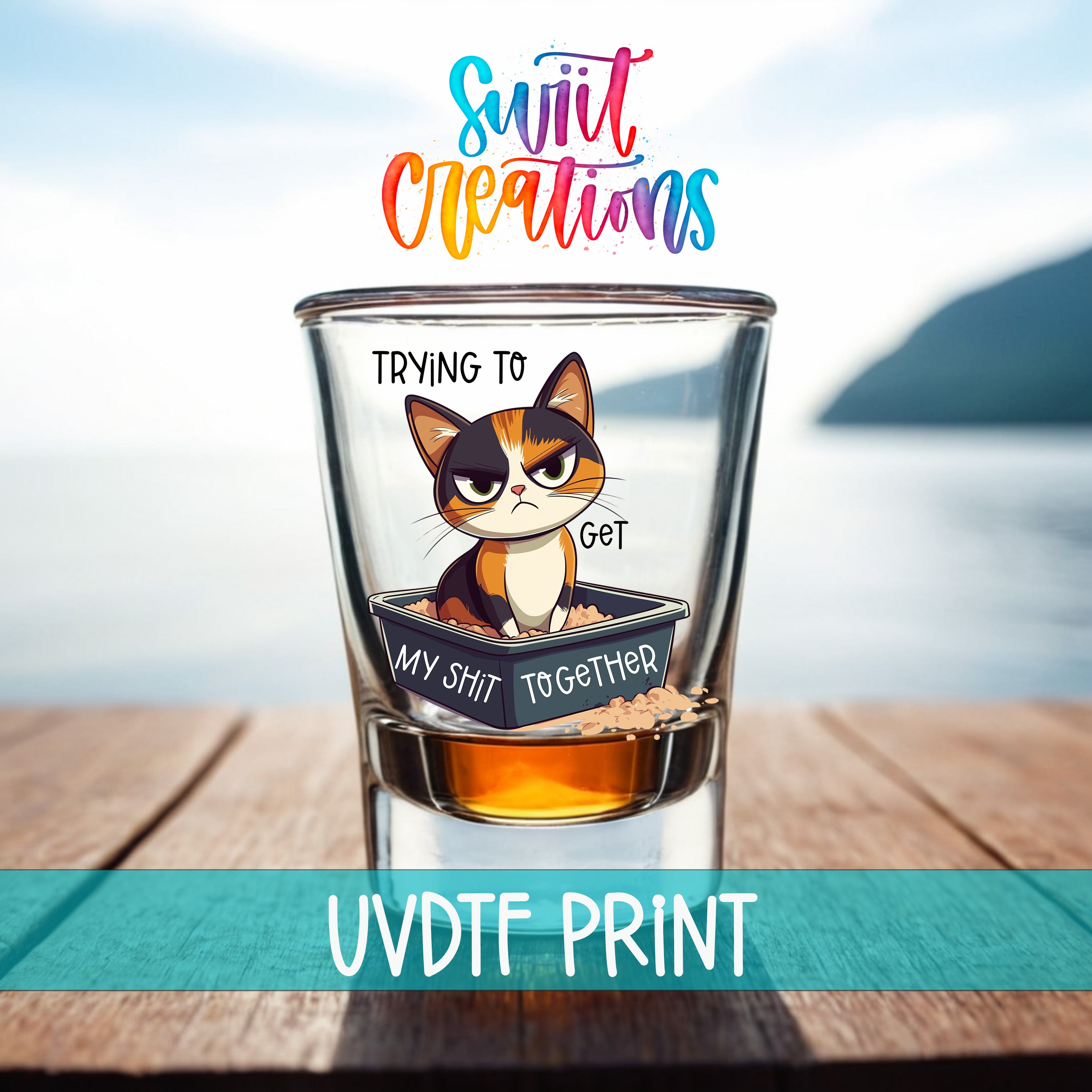 A glass with a cartoon cat and the text "Trying to get my shit together" printed on it, set against a blurred background of a body of water and a mountain.