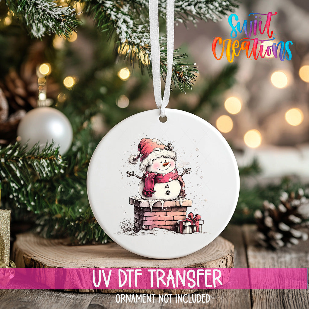 A white ceramic ornament with a snowman and presents is hanging on a Christmas tree, with a pink ribbon around it.