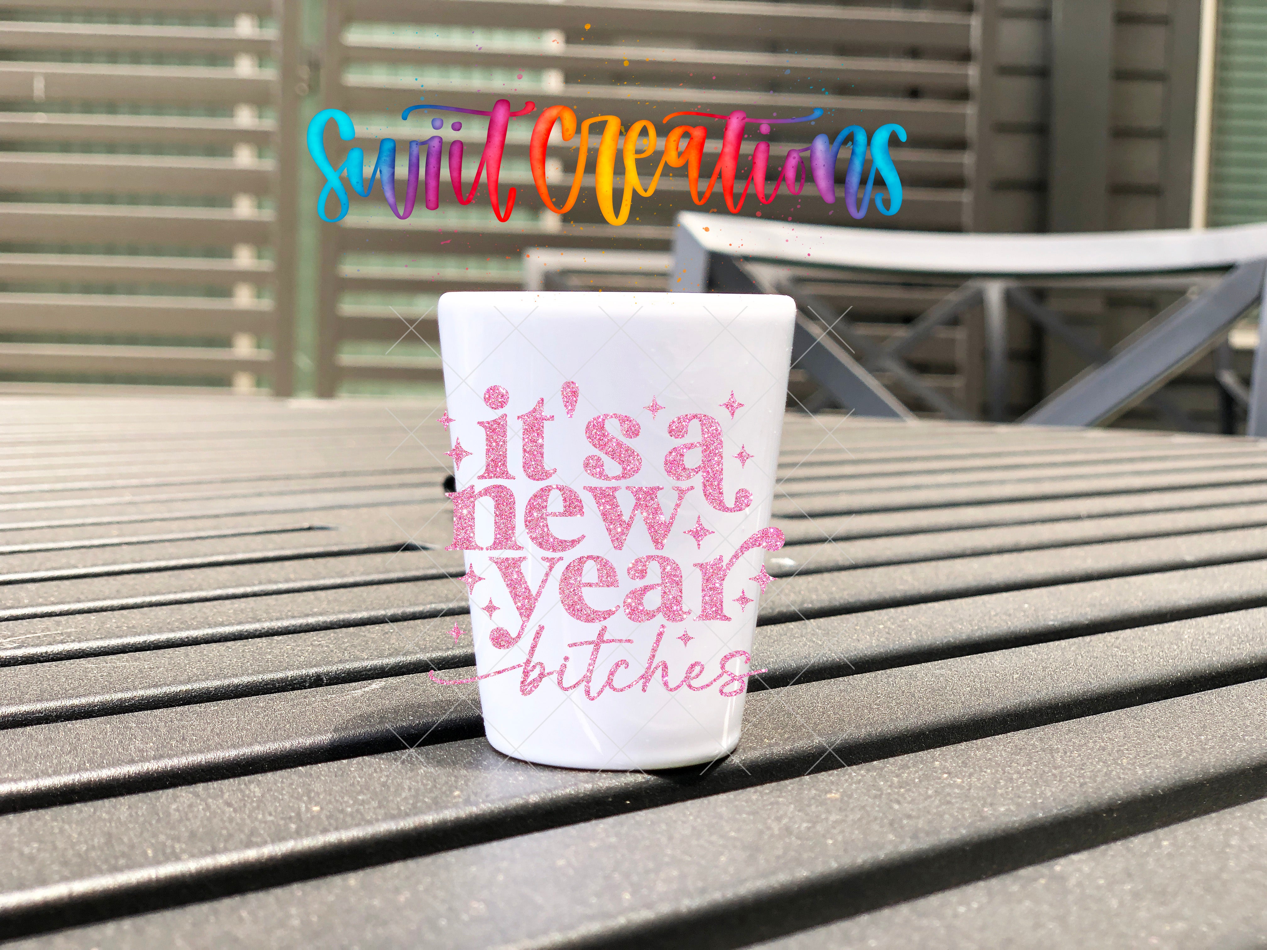 A white cup with pink text that says "it's a new year, ladies - bitches" is placed on a wooden surface.