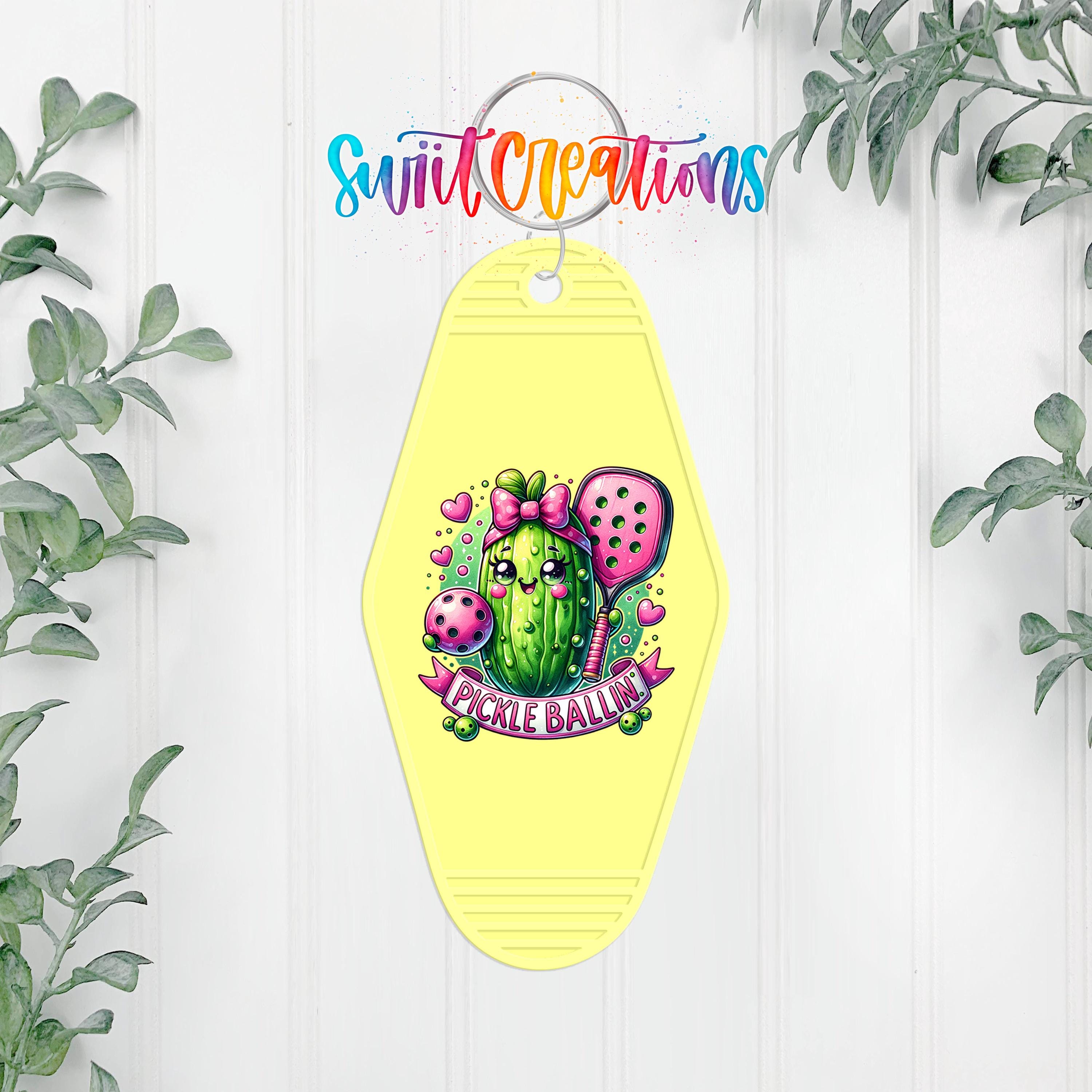 A yellow keychain with a colorful illustration of a pickle and a banner that says "Pickle Ballin'" hanging on a white wall.