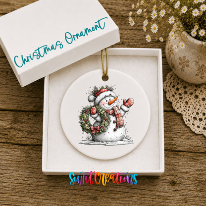 snowman in a red hat holding a Christmas ornament ceramic Christmas ornament presented in a gift box