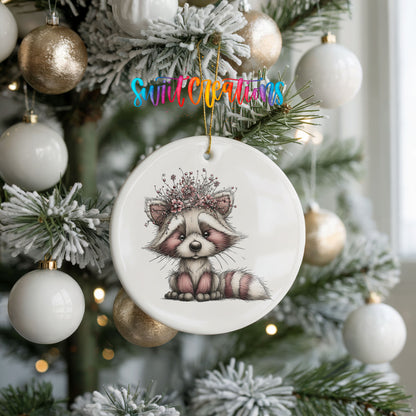 woodland raccoon wearing a pink winter bow ceramic Christmas ornament hanging on a frosted tree with ornaments