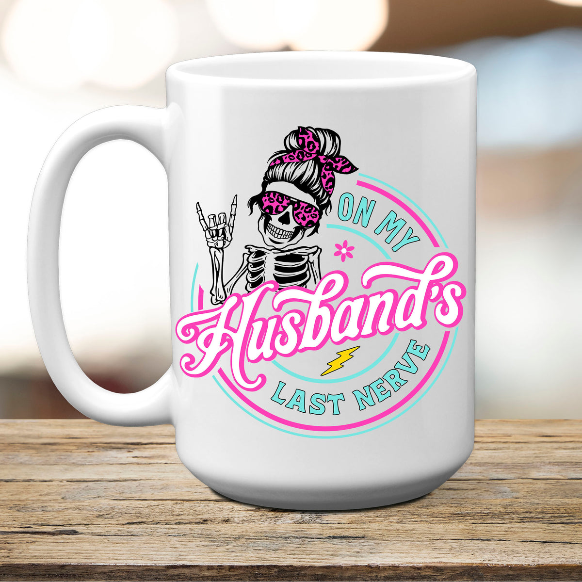 On My Husband's Last Nerve funny 15oz ceramic mug with a messy bun skeleton in pink leopard sunglasses, on a wood table