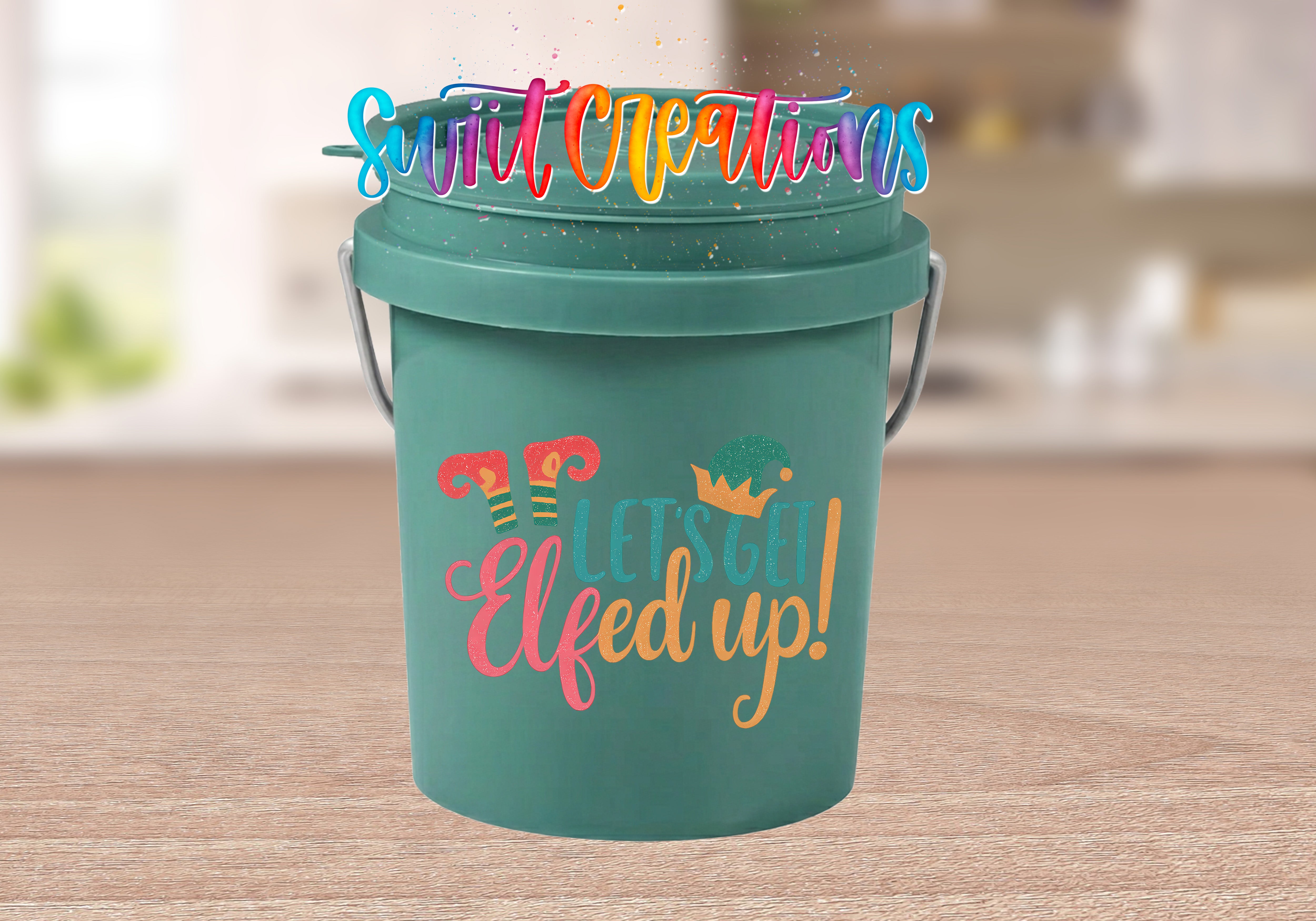Let's Get Elfed Up Mini Bucket UV DTF Transfer with coral, teal, and golden orange lettering, striped elf legs, and an elf hat on a teal mini bucket displayed on a wood tabletop