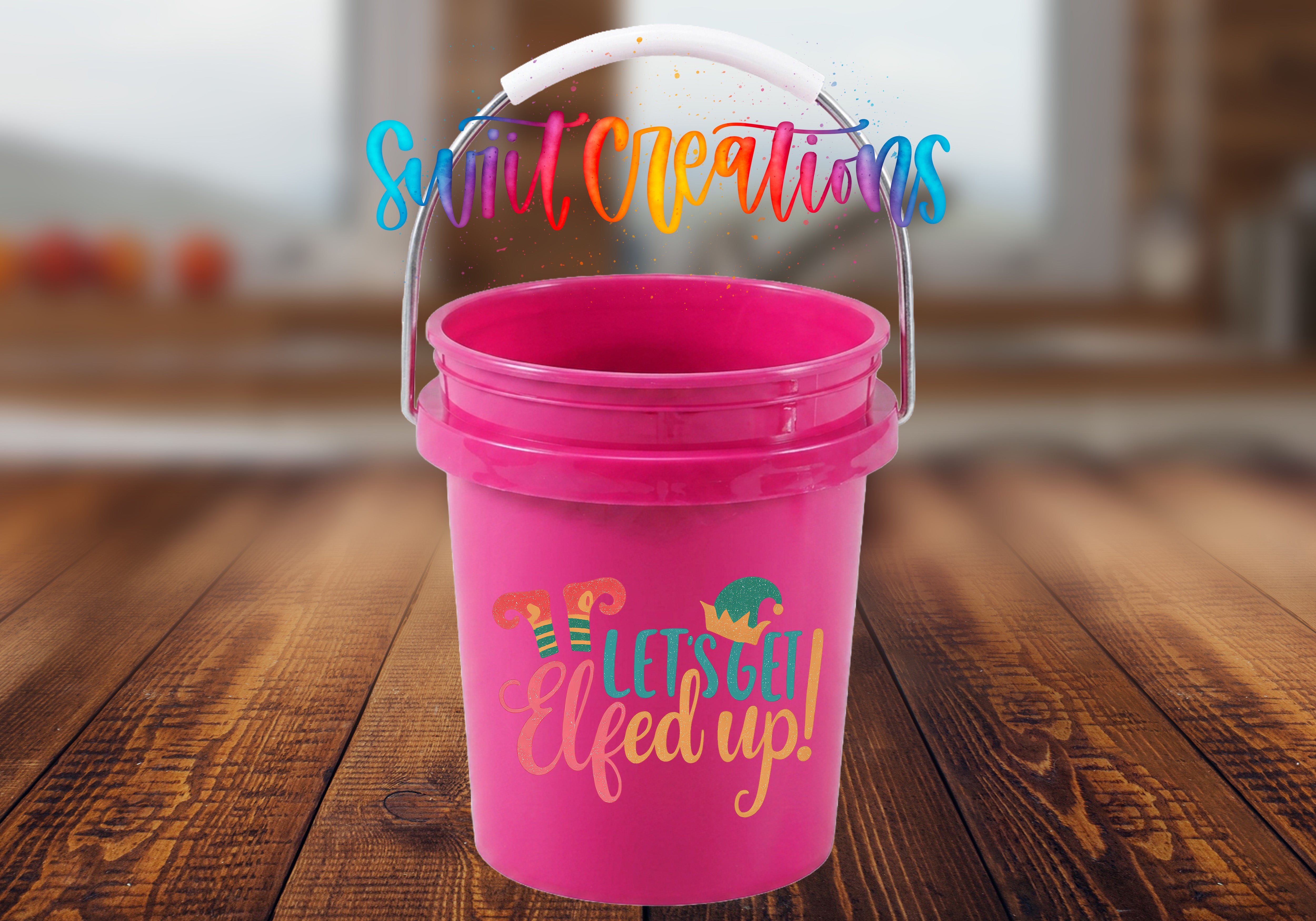 Let's Get Elfed Up Mini Bucket UV DTF Transfer with coral, teal, and golden orange lettering, striped elf legs, and an elf hat on a pink mini bucket with handle raised on a rustic wood tabletop