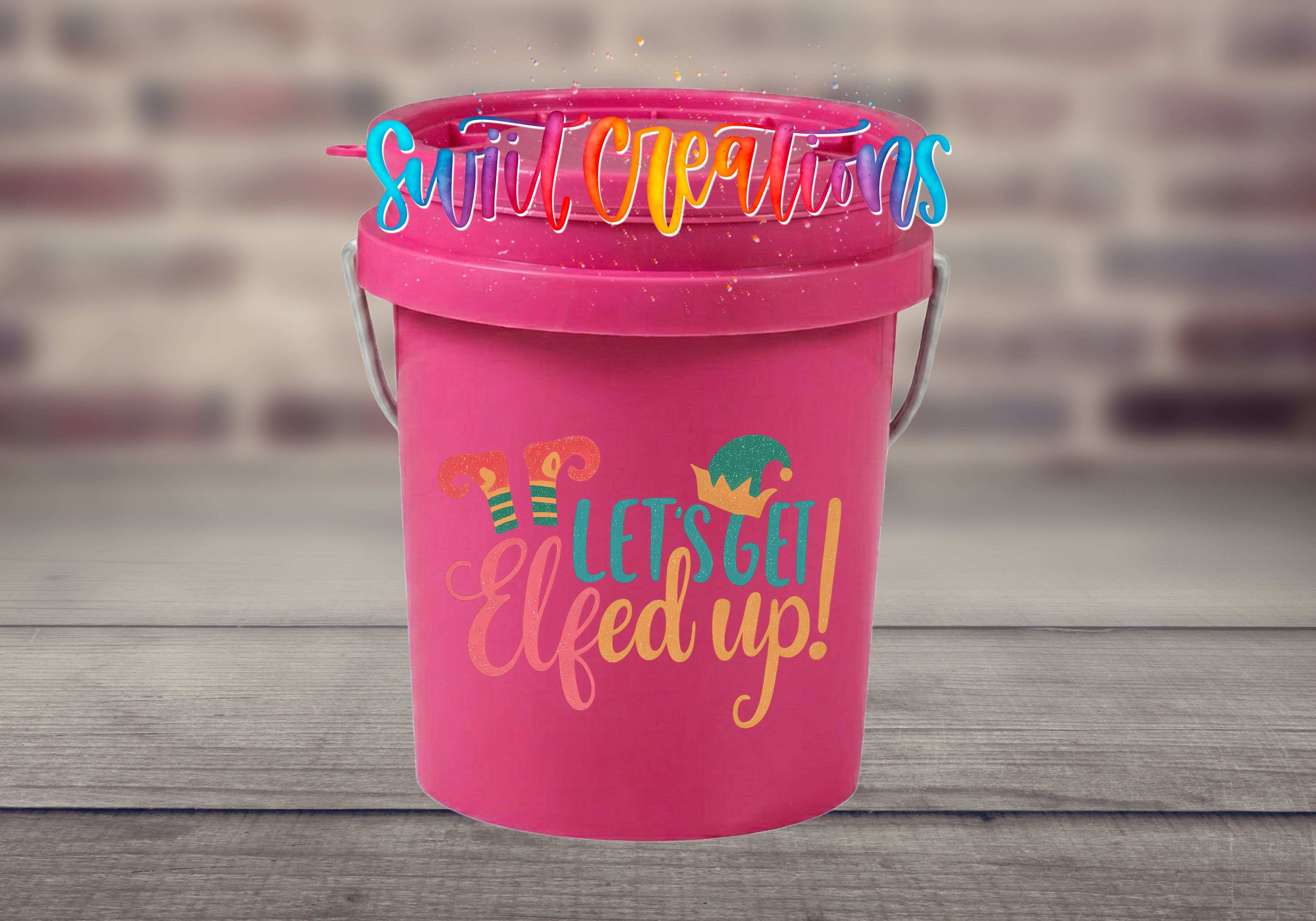 Let's Get Elfed Up Mini Bucket UV DTF Transfer with coral, teal, and golden orange lettering, striped elf legs, and an elf hat on a pink mini bucket displayed on a gray wood surface