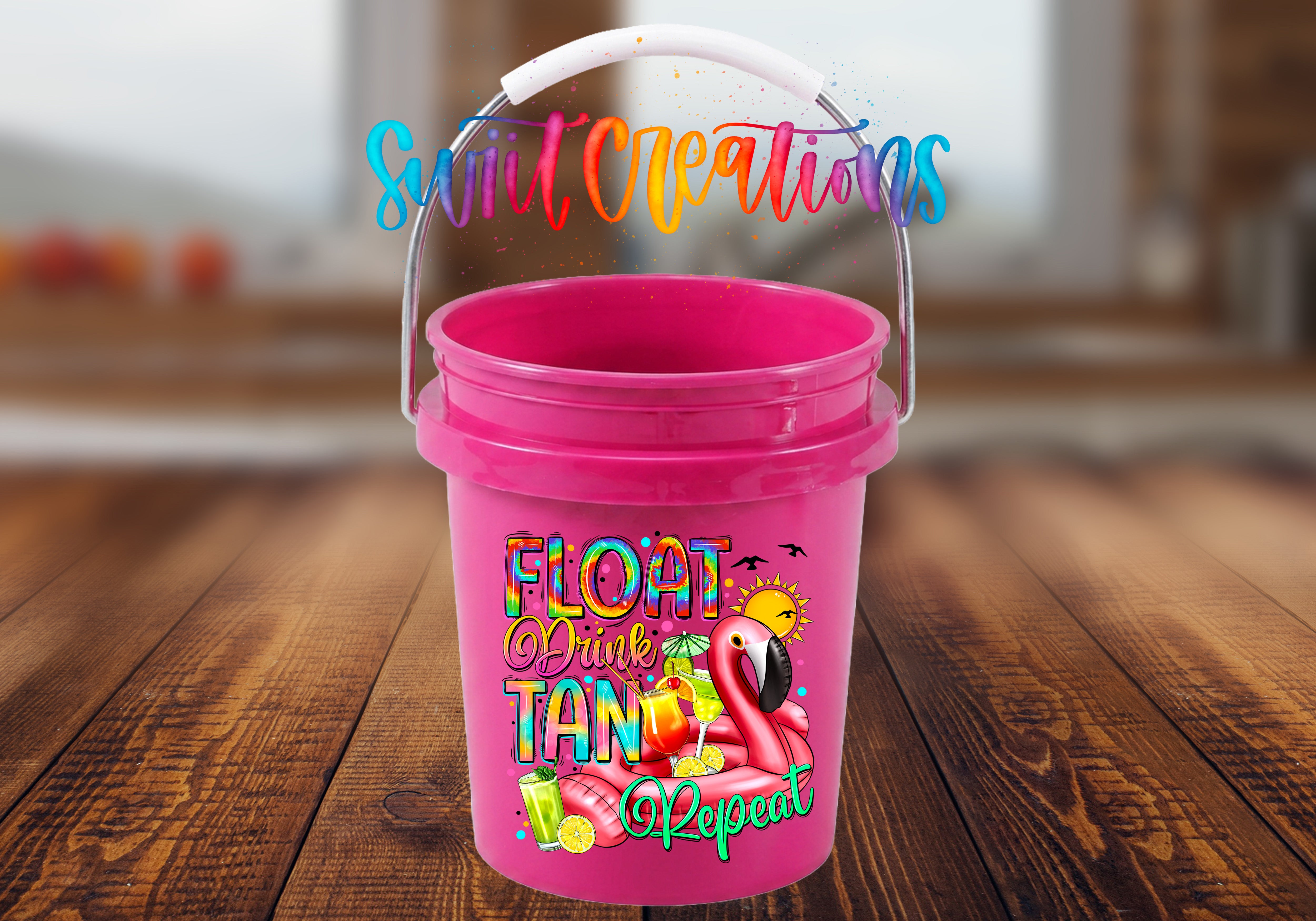 Float Drink Tan Repeat Mini Bucket UV DTF Transfer with rainbow lettering, pink flamingo pool float, tropical cocktails, citrus, sun, and birds on a pink mini bucket with handle raised on a rustic wood tabletop