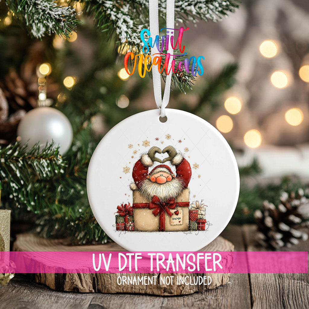 Cute Santa in a gift box ornament UV DTF transfer on an ornament resting on a wood slice beside a Christmas tree