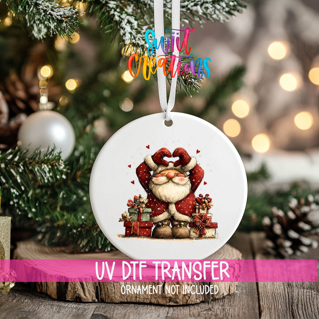 Cute Santa making a heart over his head with gifts ornament UV DTF transfer on an ornament resting on a wood slice beside a Christmas tree