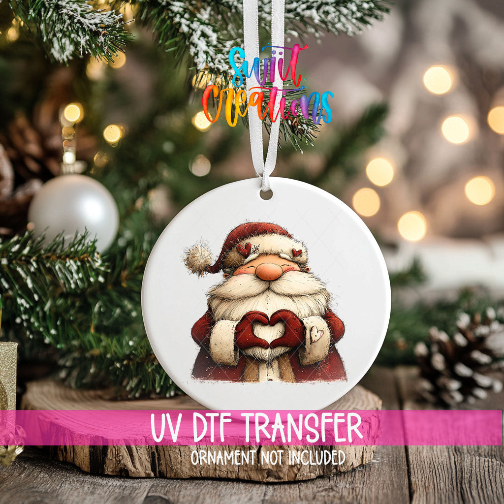 Cute Santa making heart hands ornament UV DTF transfer on an ornament resting on a wood slice beside a Christmas tree