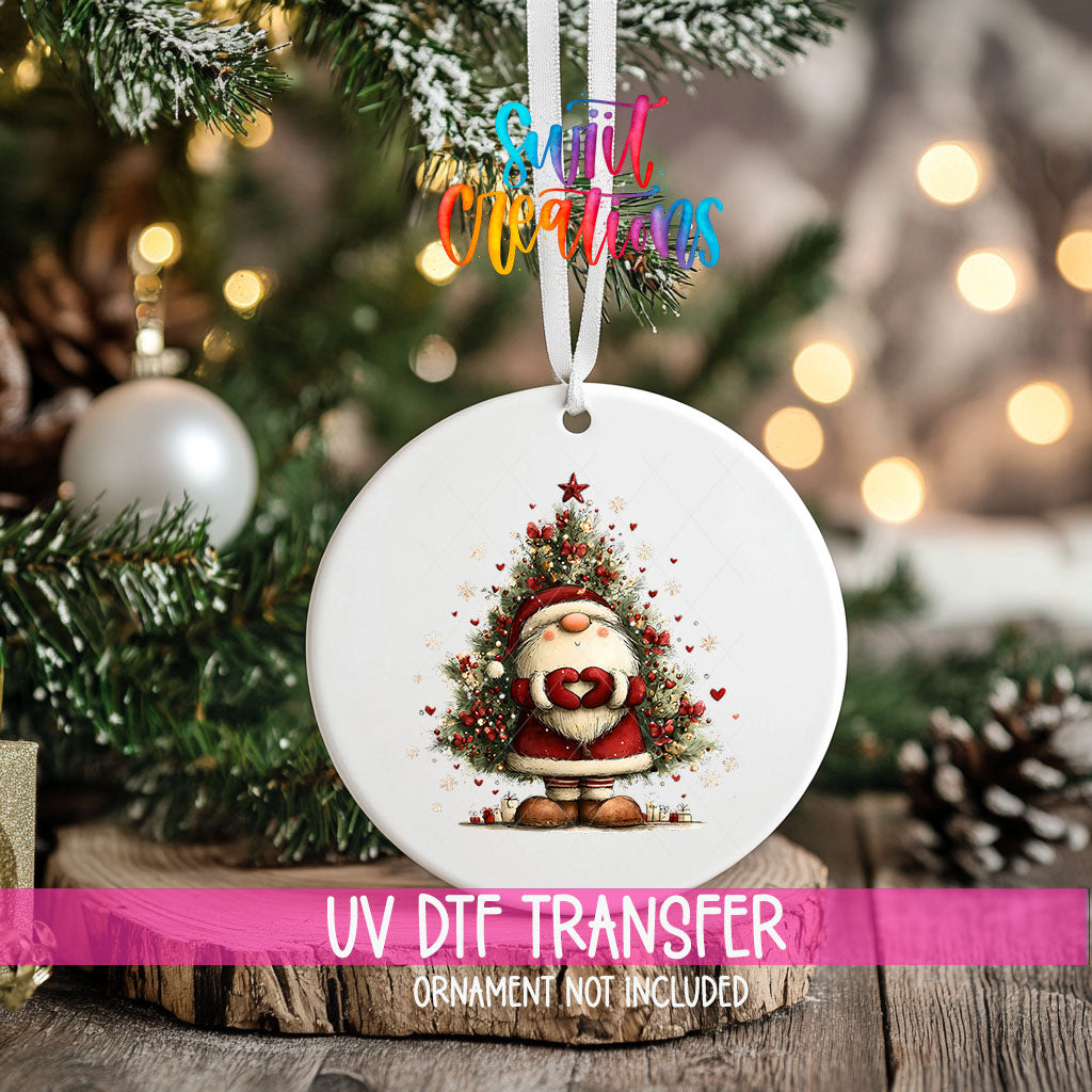 Cute Santa with heart hands in front of a Christmas tree ornament UV DTF transfer on an ornament resting on a wood slice beside a Christmas tree