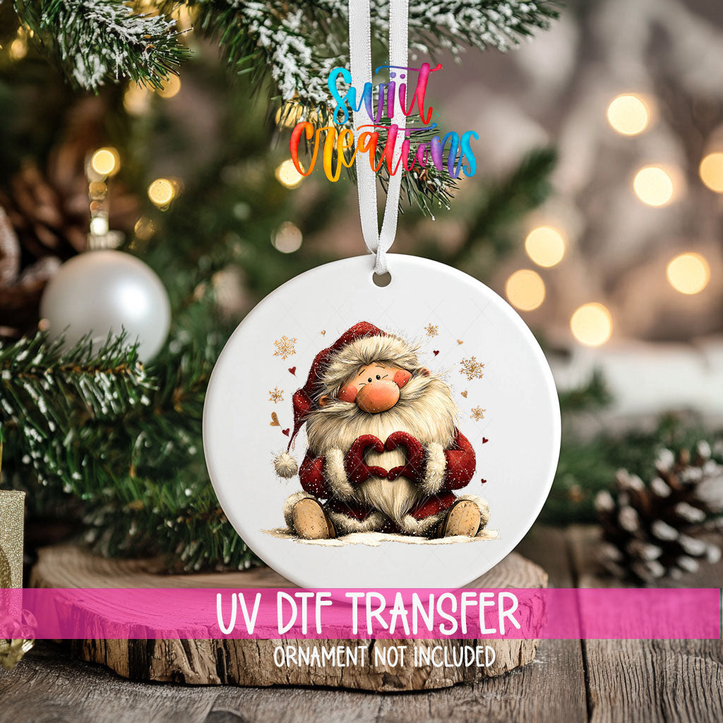 Cute sitting Santa making heart hands ornament UV DTF transfer on an ornament resting on a wood slice beside a Christmas tree