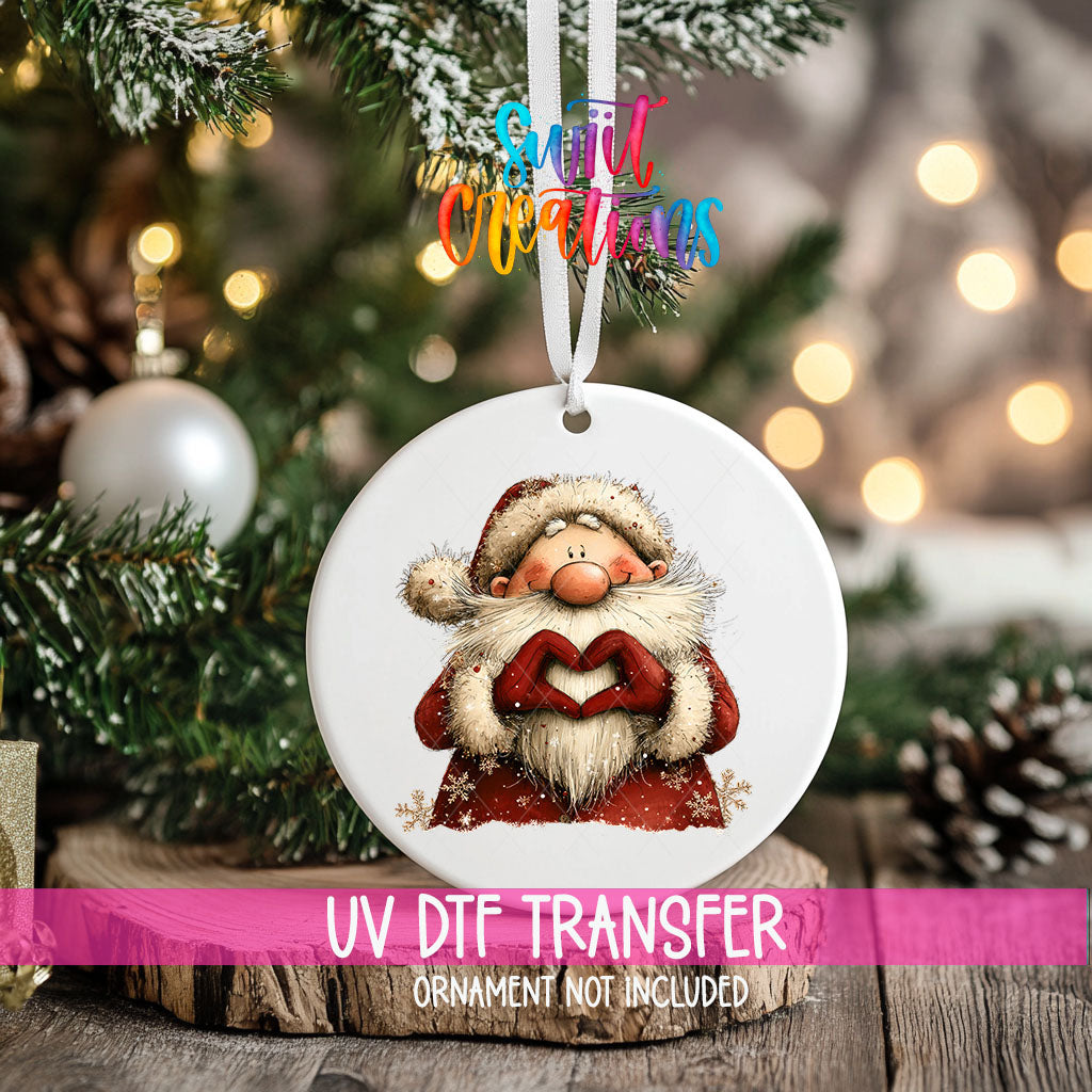 Cute Santa in a snowflake coat making heart hands ornament UV DTF transfer on an ornament resting on a wood slice beside a Christmas tree