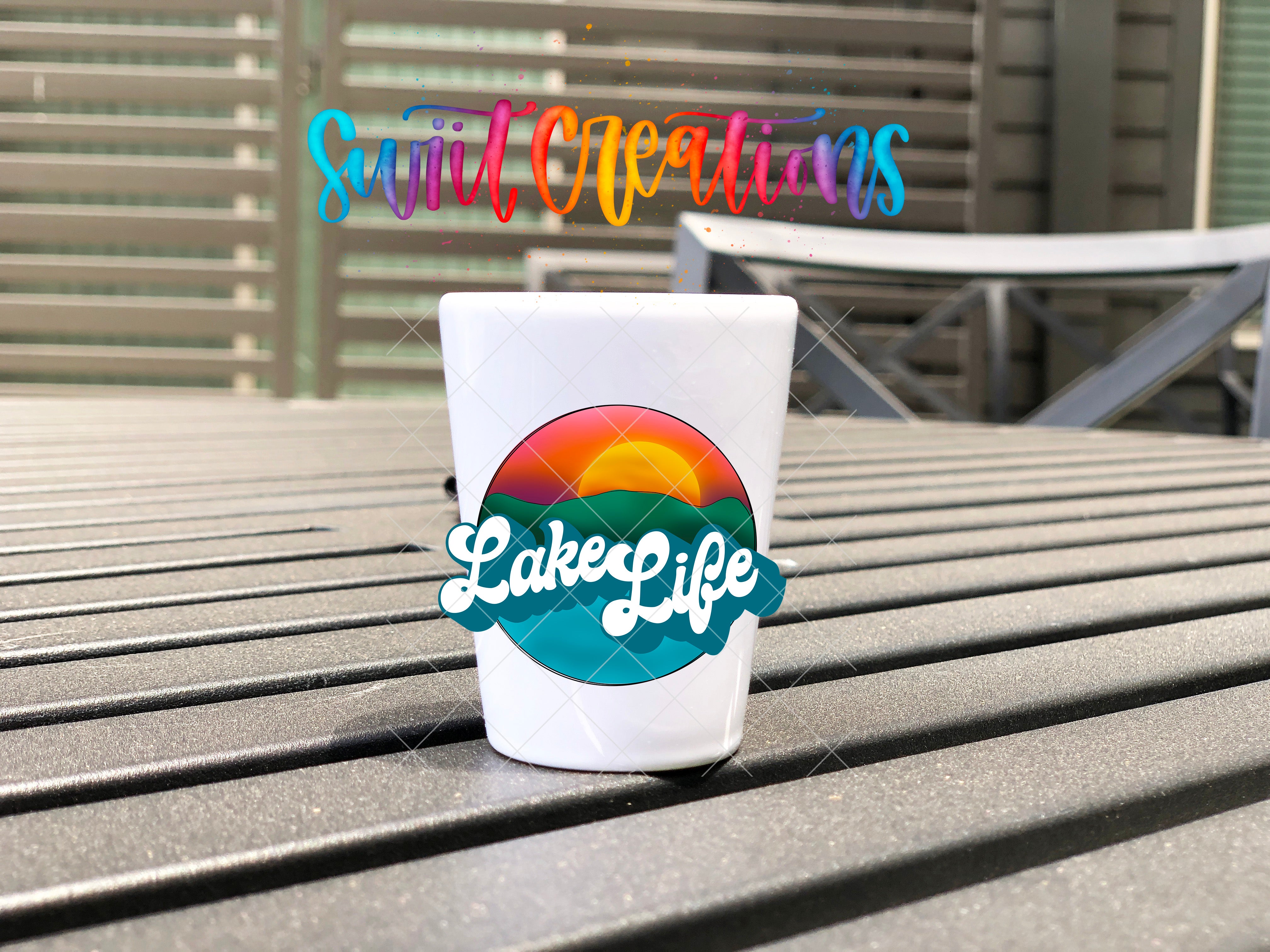 A white shot glass with a colorful design and the text "LakesLife" on it, placed on a wooden surface.
