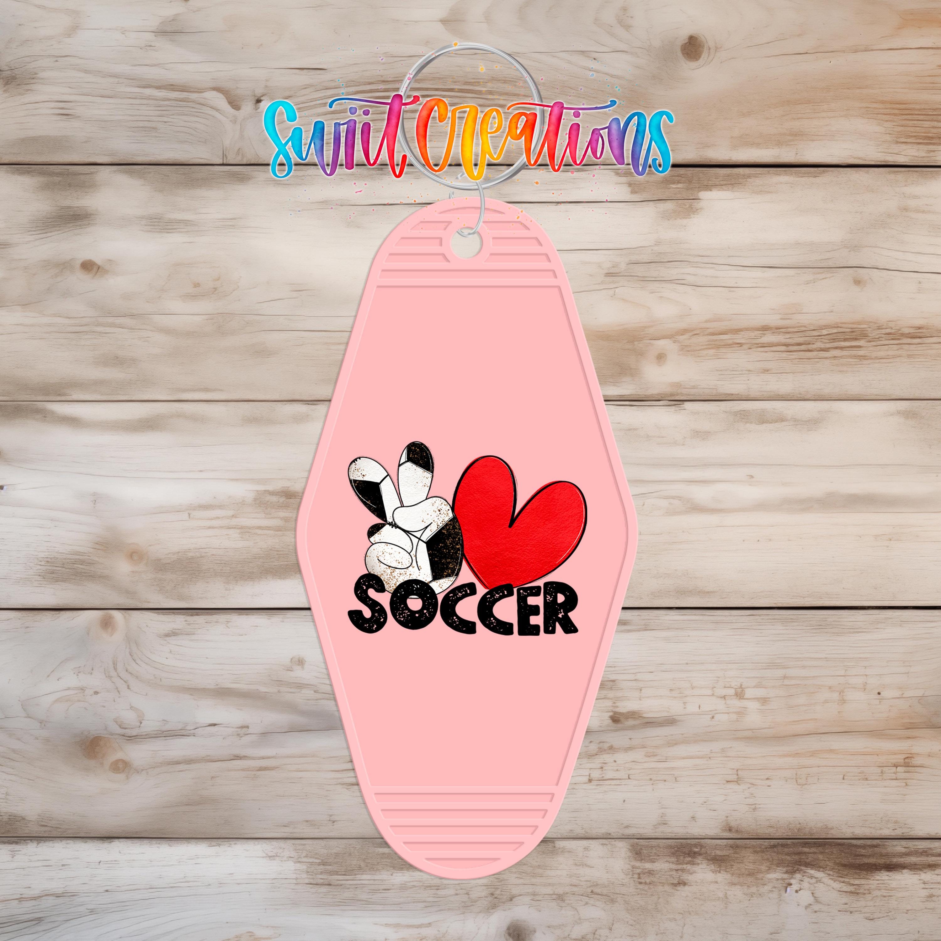 A pink keychain with a soccer ball and a heart design, along with the word "SOCCER" written on it, is displayed on a wooden surface.
